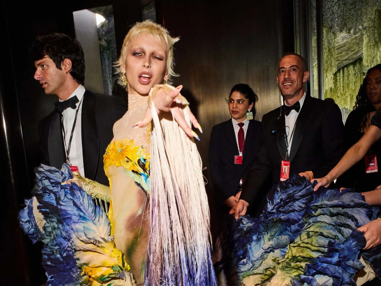 Center-stage blonde woman in a beige fringed dress points at the camera while surrounded by suited staff with lanyards on backstage at a fashion show.