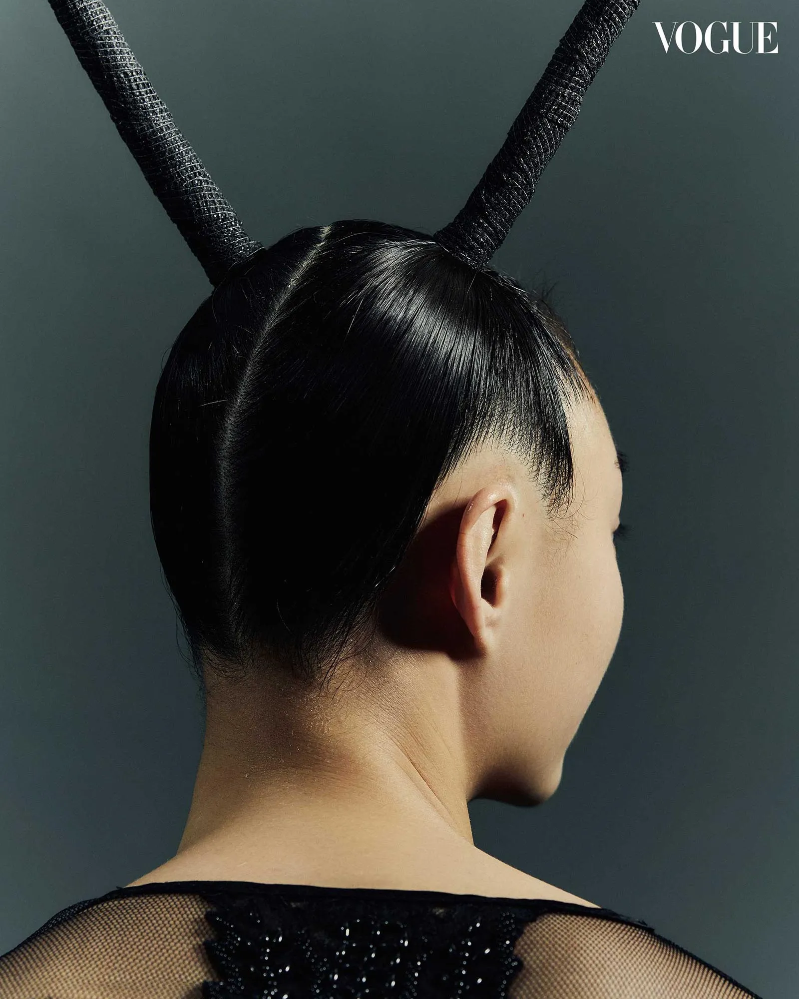 Back view of a model wearing two large black horn-like headpieces against a dark backdrop, Vogue logo in corner.