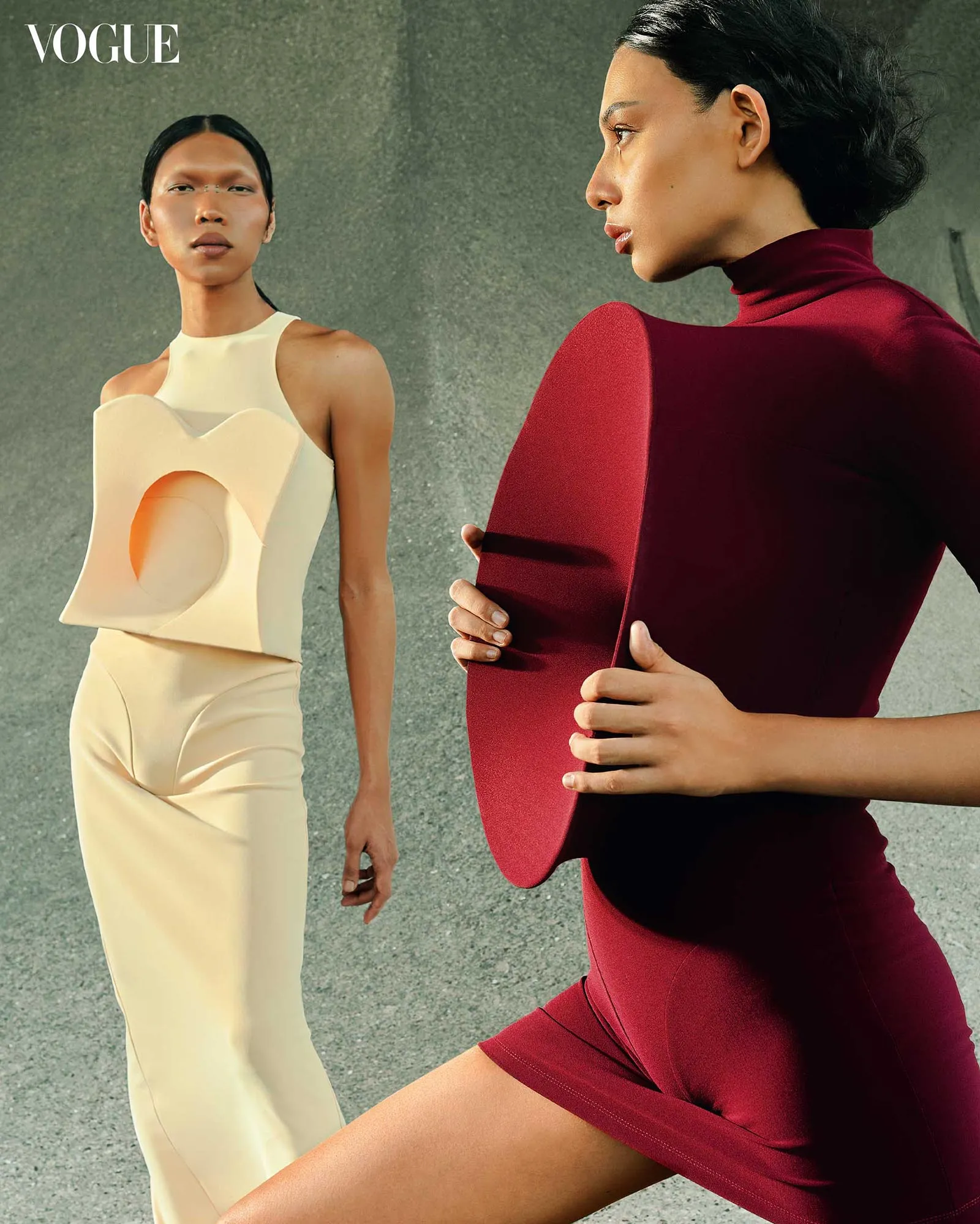 Editorial fashion shot for Vogue: two models in sculptural dresses against a gray concrete backdrop, one in a cream geometric gown and the other in a red burgundy form-fitting outfit holding a circular piece.
