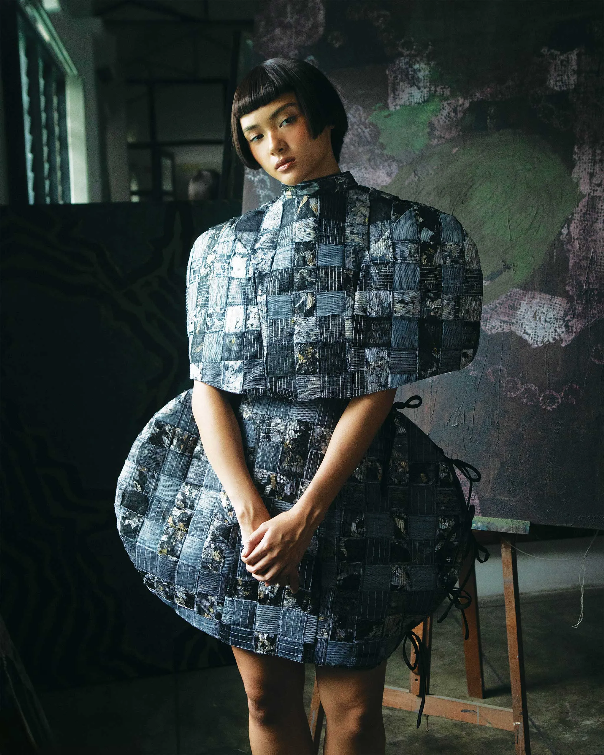 Woman with short dark hair wearing a blue patchwork quilted dress, standing in a dim studio.