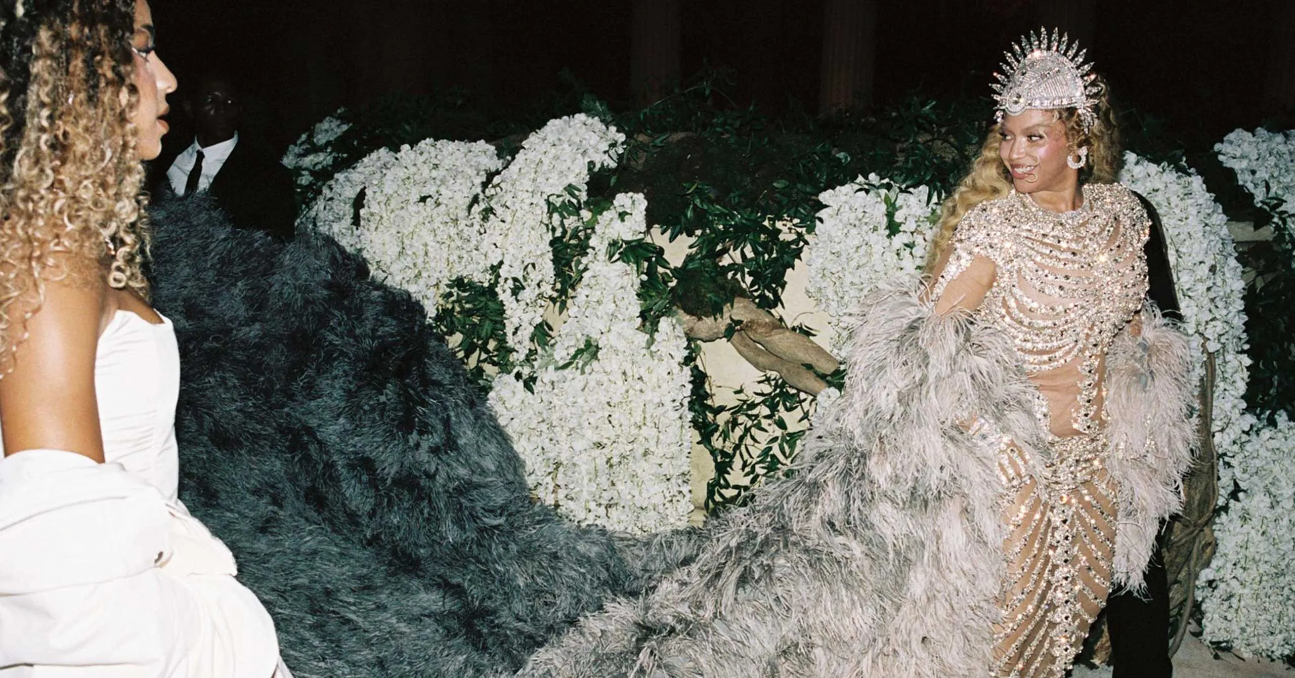 Woman in a sparkling jeweled gown and crown smiles, standing near a backdrop of white flowers; another woman in a white dress is nearby.