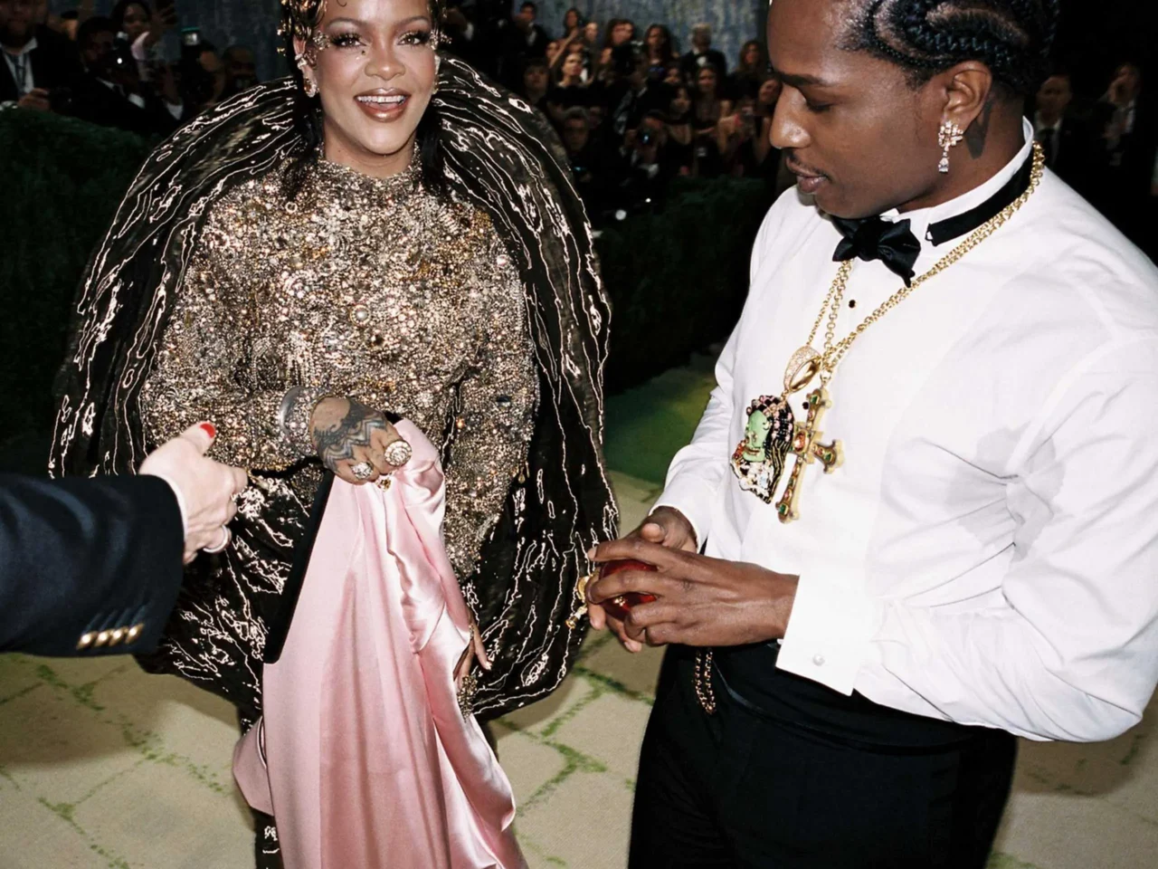 Woman in a sparkling jeweled gown and crown smiles, standing near a backdrop of white flowers; another woman in a white dress is nearby.