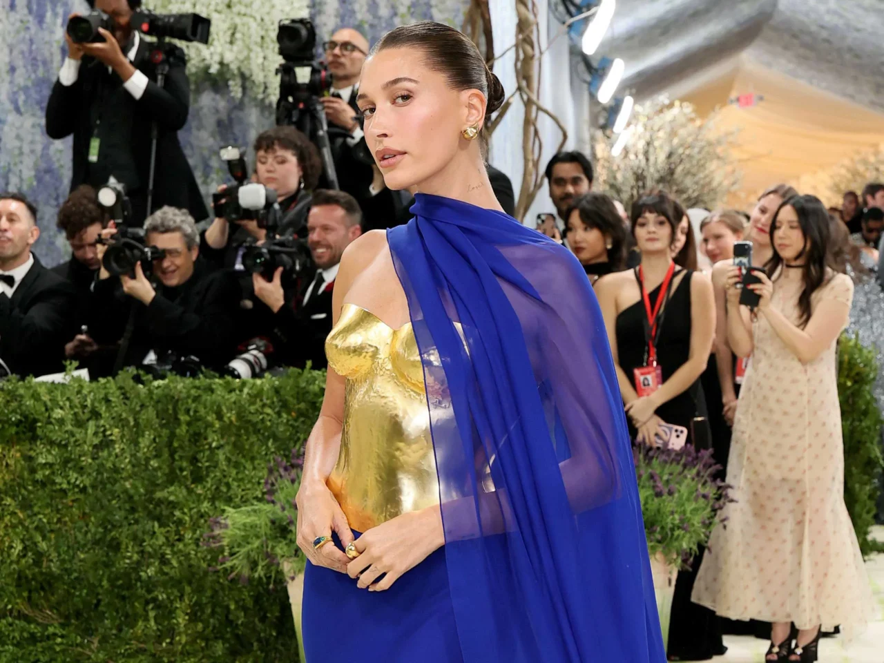 Woman in a gold metallic dress with a blue sheer cape posing on the red carpet as photographers surround her, capturing the moment.