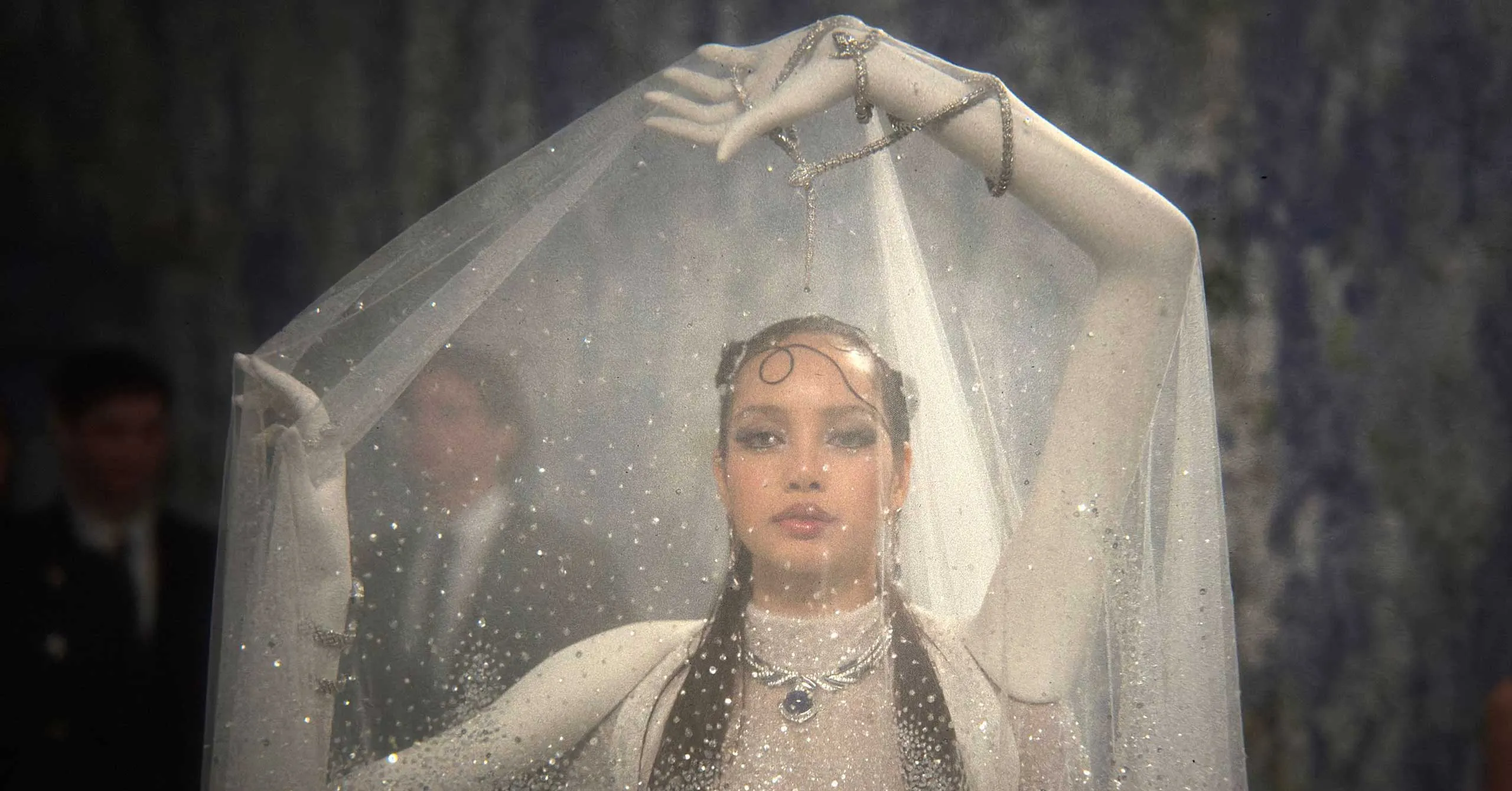 Bride wearing a sheer veil and glittering jewelry, standing outdoors with rain droplets on the veil