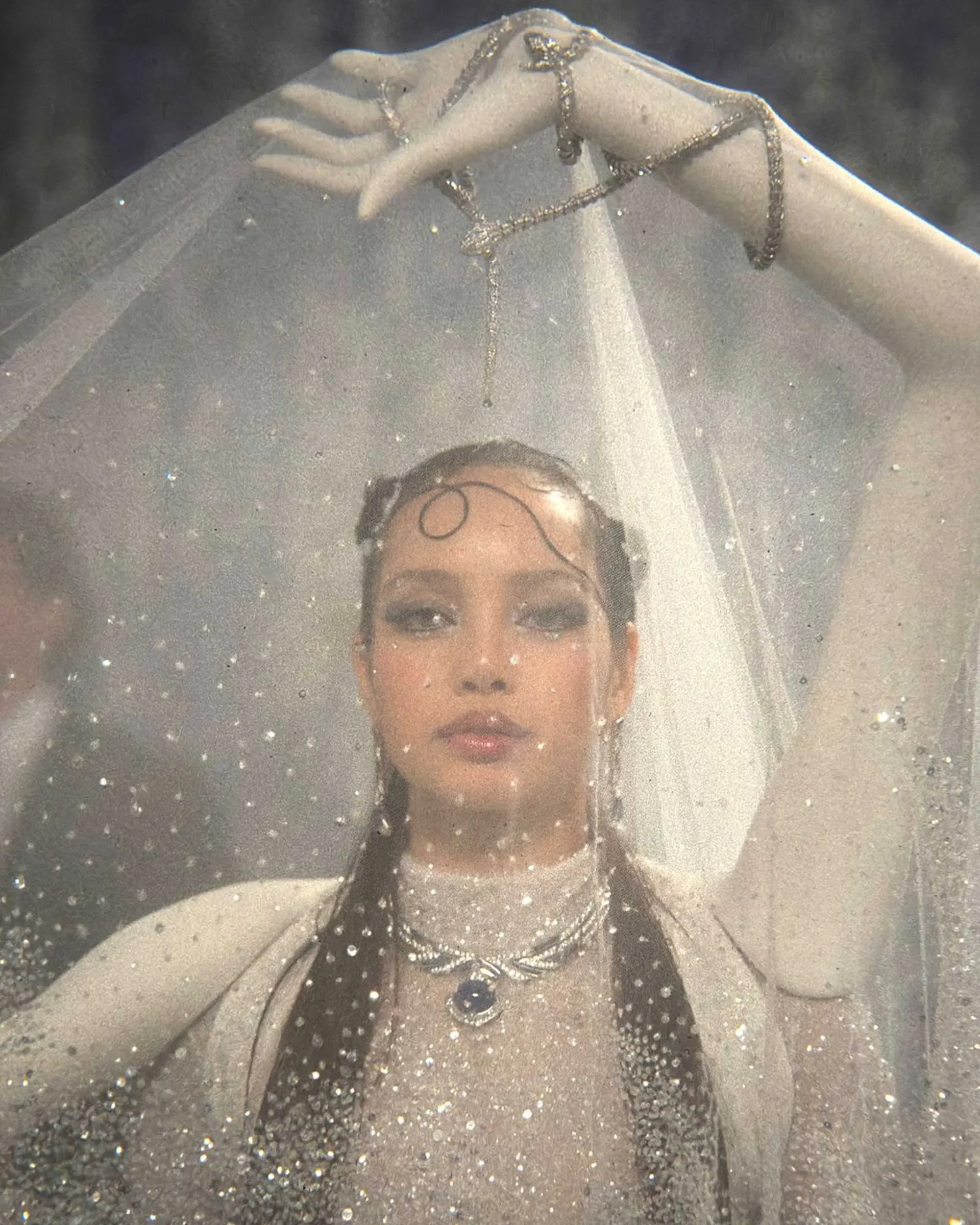 Bride wearing a sheer veil and glittering jewelry, standing outdoors with rain droplets on the veil