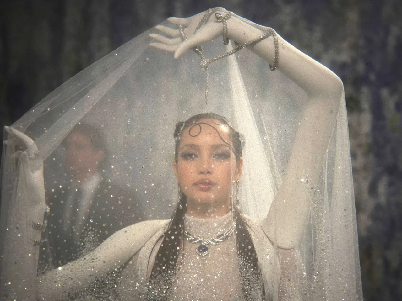 Bride wearing a sheer veil and glittering jewelry, standing outdoors with rain droplets on the veil