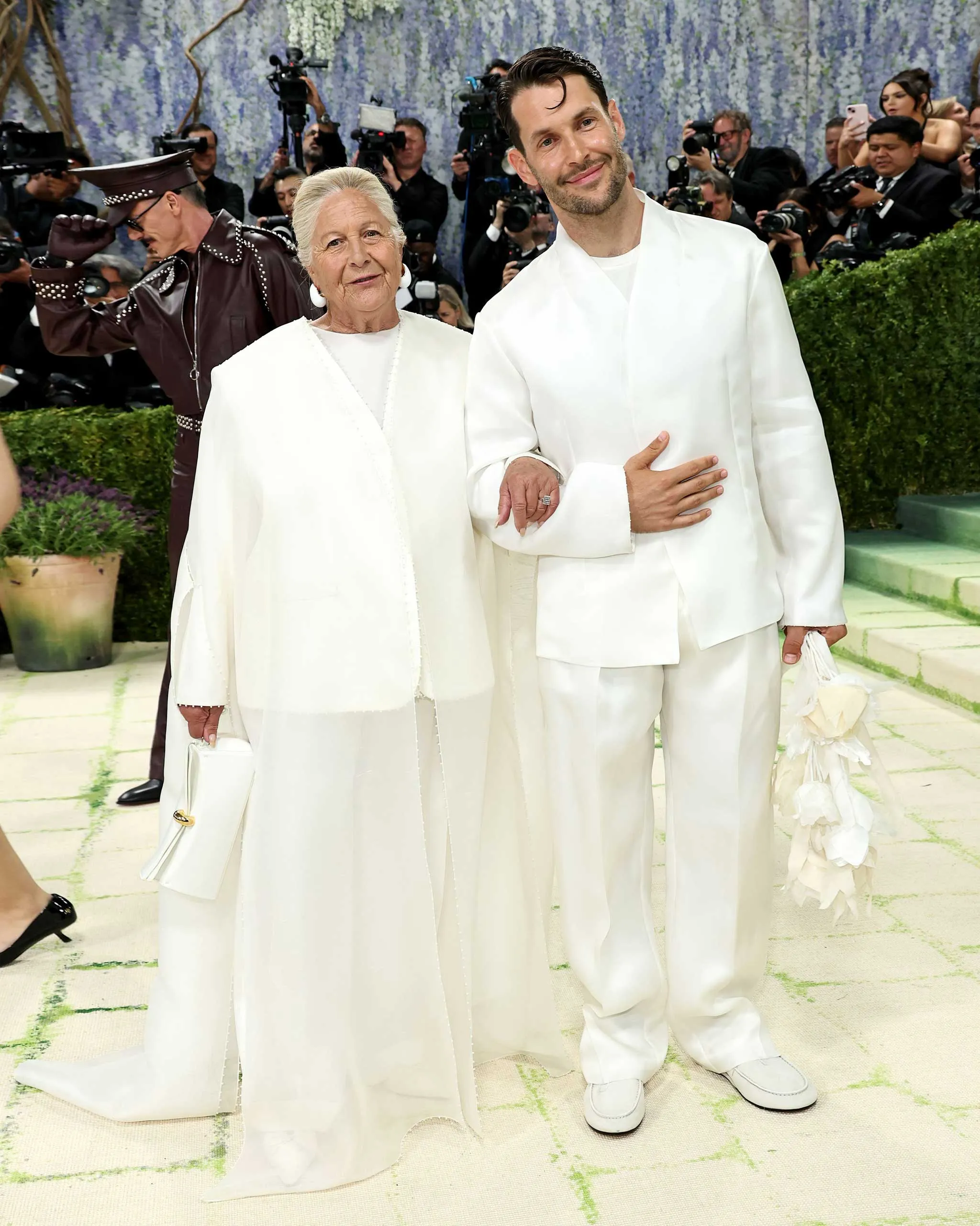Older woman and younger man in white outfits posing on a red-carpet event as paparazzi flash behind them