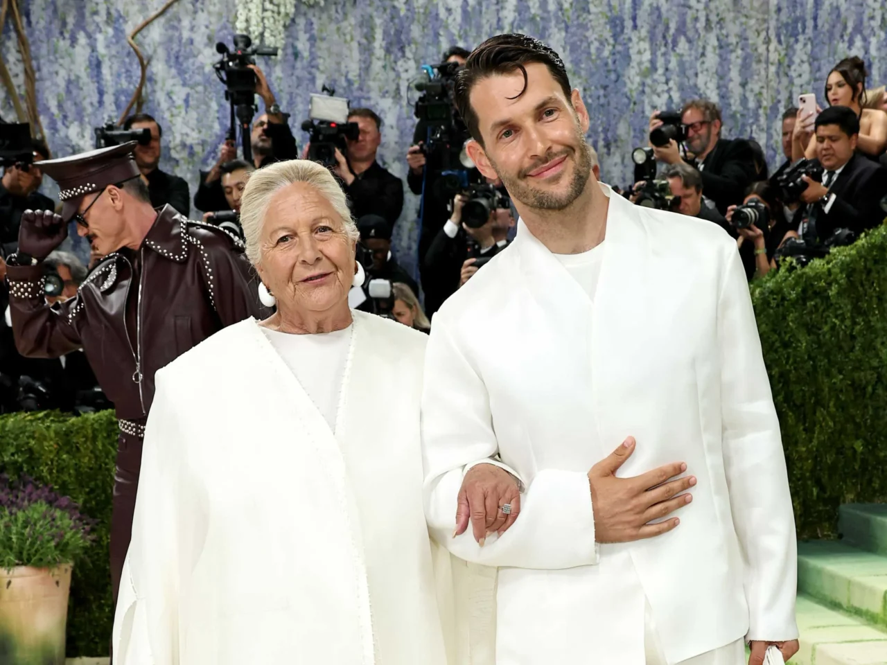 Two people in white outfits pose together on a red carpet as photographers swarm in the background against a floral backdrop.
