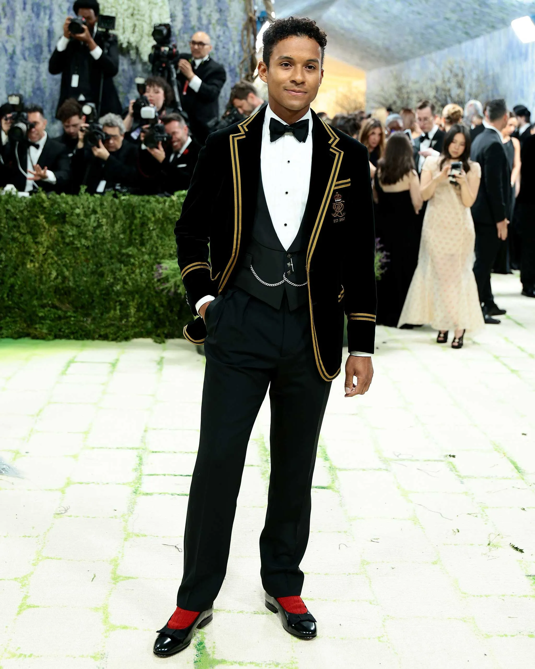 Young man in a black velvet tuxedo with gold piping and bow tie, posing on a gala carpet as photographers snap in the background.