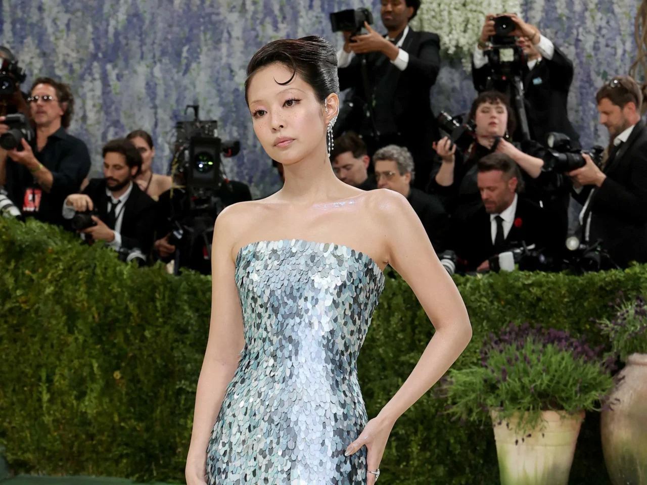 Woman in a pale blue sequined strapless gown posing on a red carpet as photographers surround her with cameras in the background.