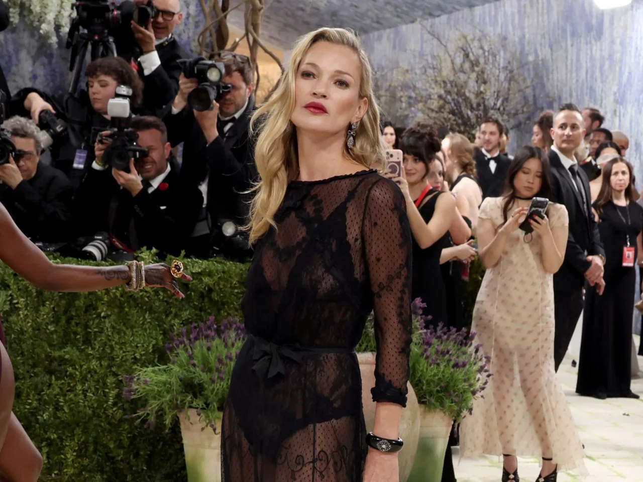 Blonde woman in a black lace dress poses on a red carpet as paparazzi photograph her.