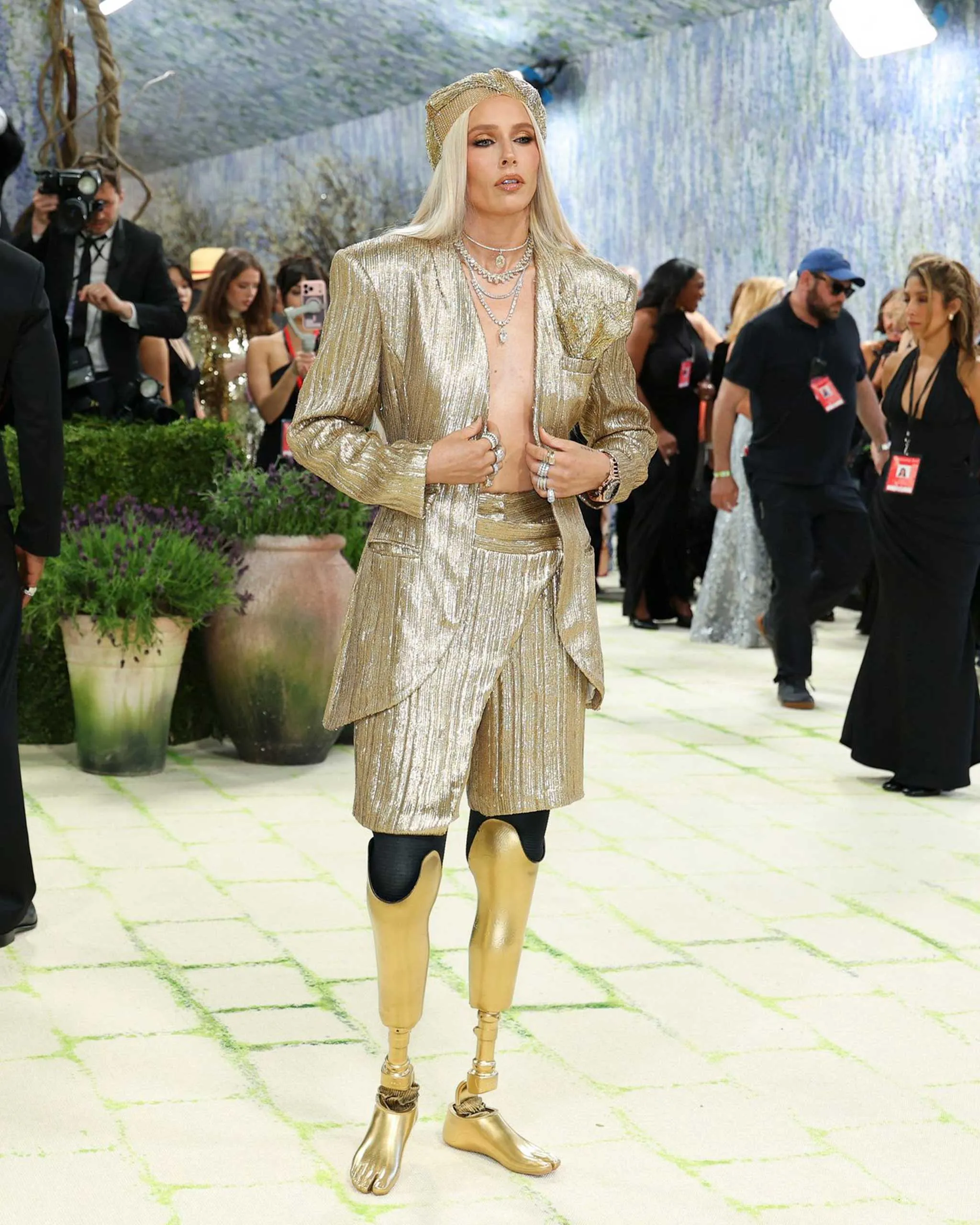 Person in a shimmering gold suit and headpiece posing on a runway with photographers and guests in the background.
