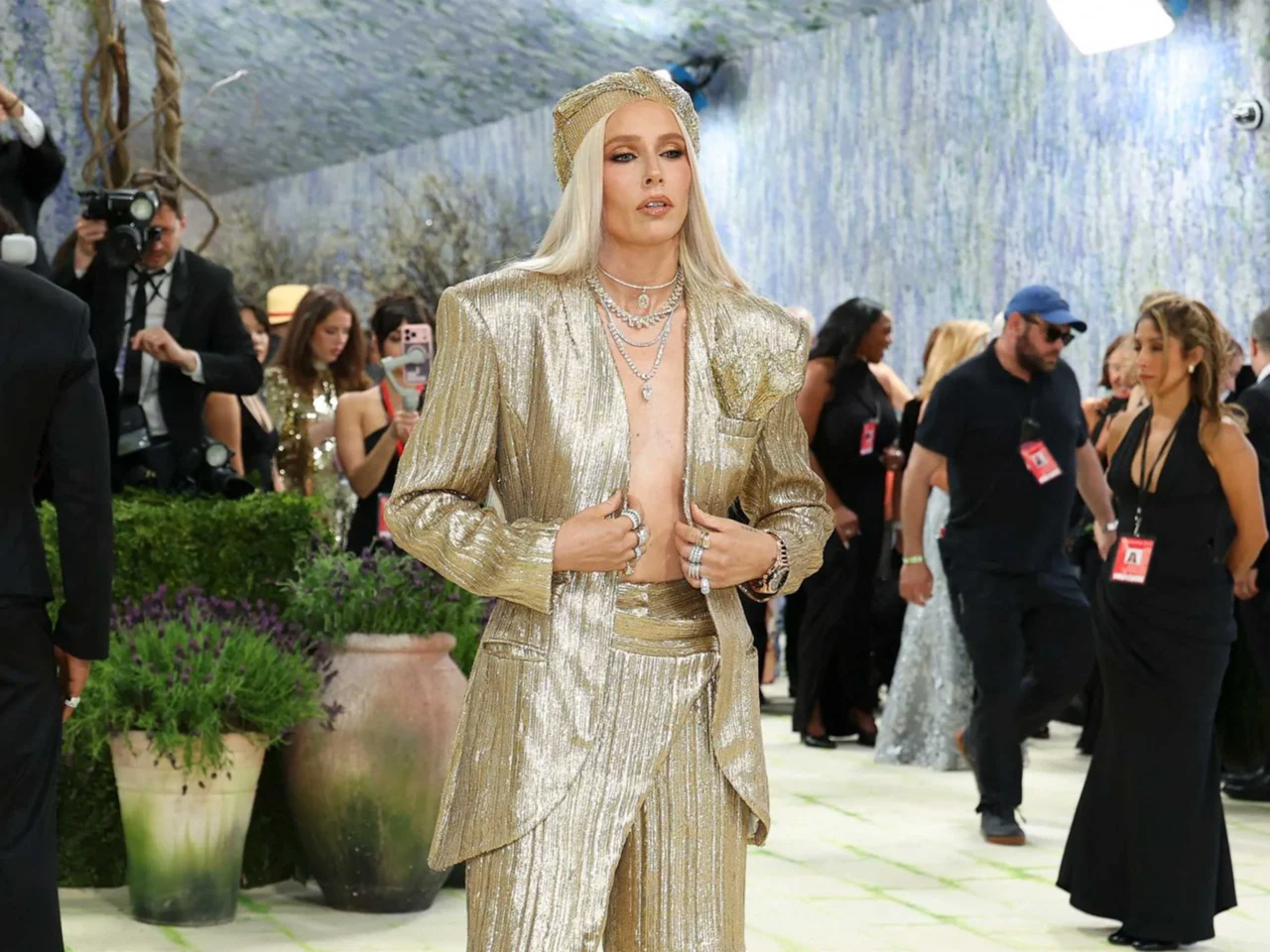 Person wearing a shimmering gold metallic suit posing on a green carpet while photographers photograph in the background