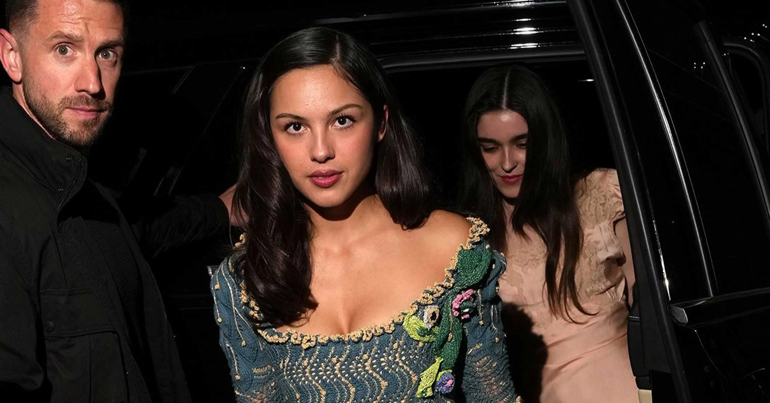 Three people exit a black car at night; center woman wears an embroidered off-shoulder dress, flanked by a man on the left and a woman on the right.