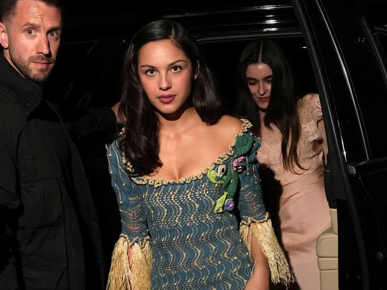 Three people exit a black car at night; center woman wears an embroidered off-shoulder dress, flanked by a man on the left and a woman on the right.