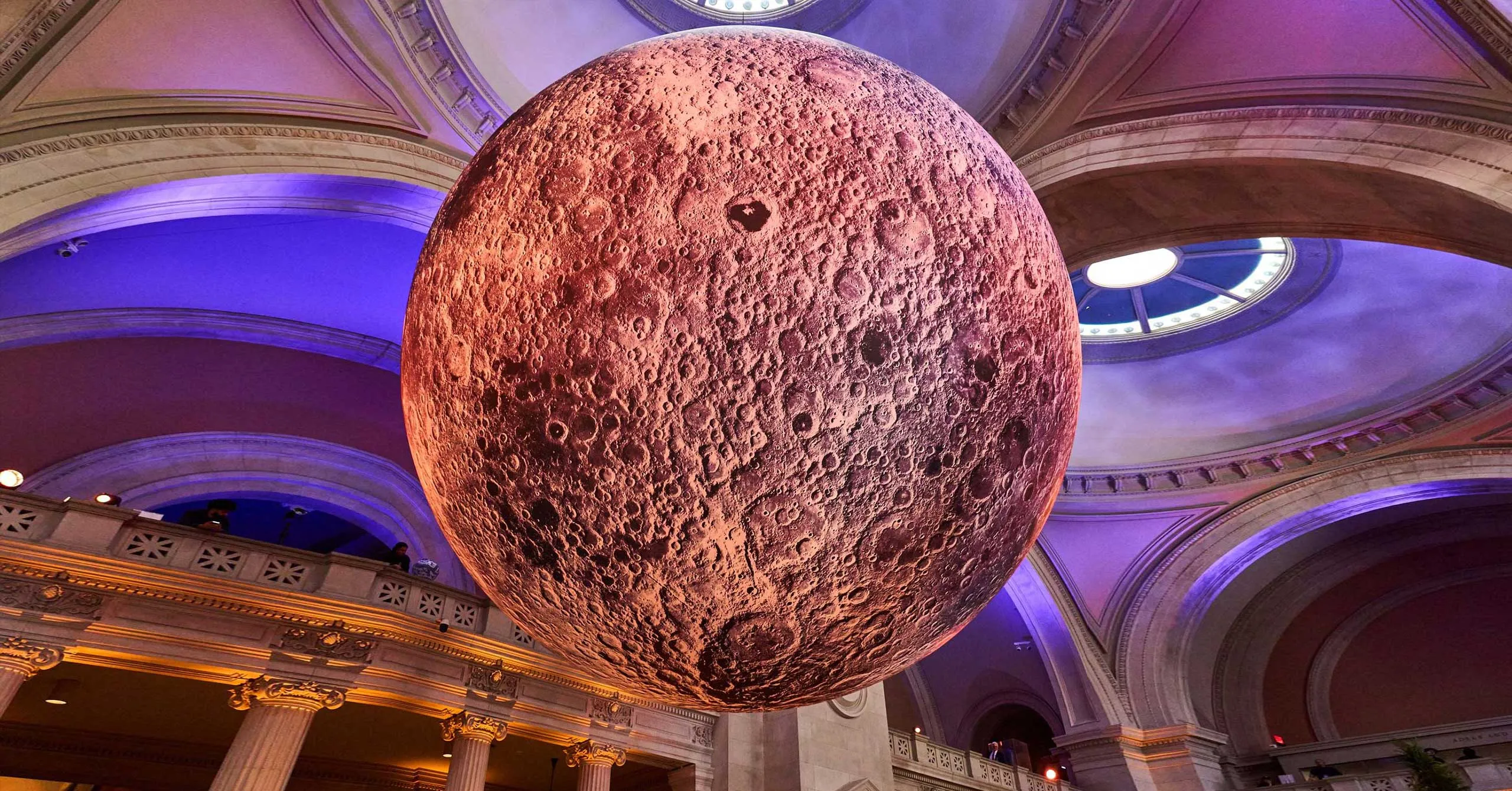 Large textured moon sculpture suspended in a grand neoclassical hall with purple lighting overhead