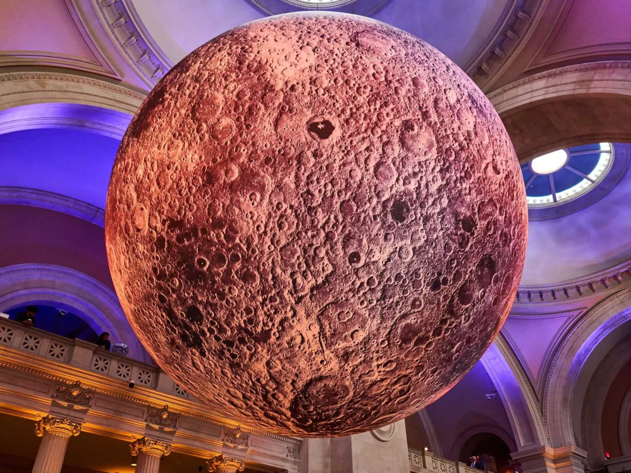 Large textured moon sculpture suspended in a grand neoclassical hall with purple lighting overhead