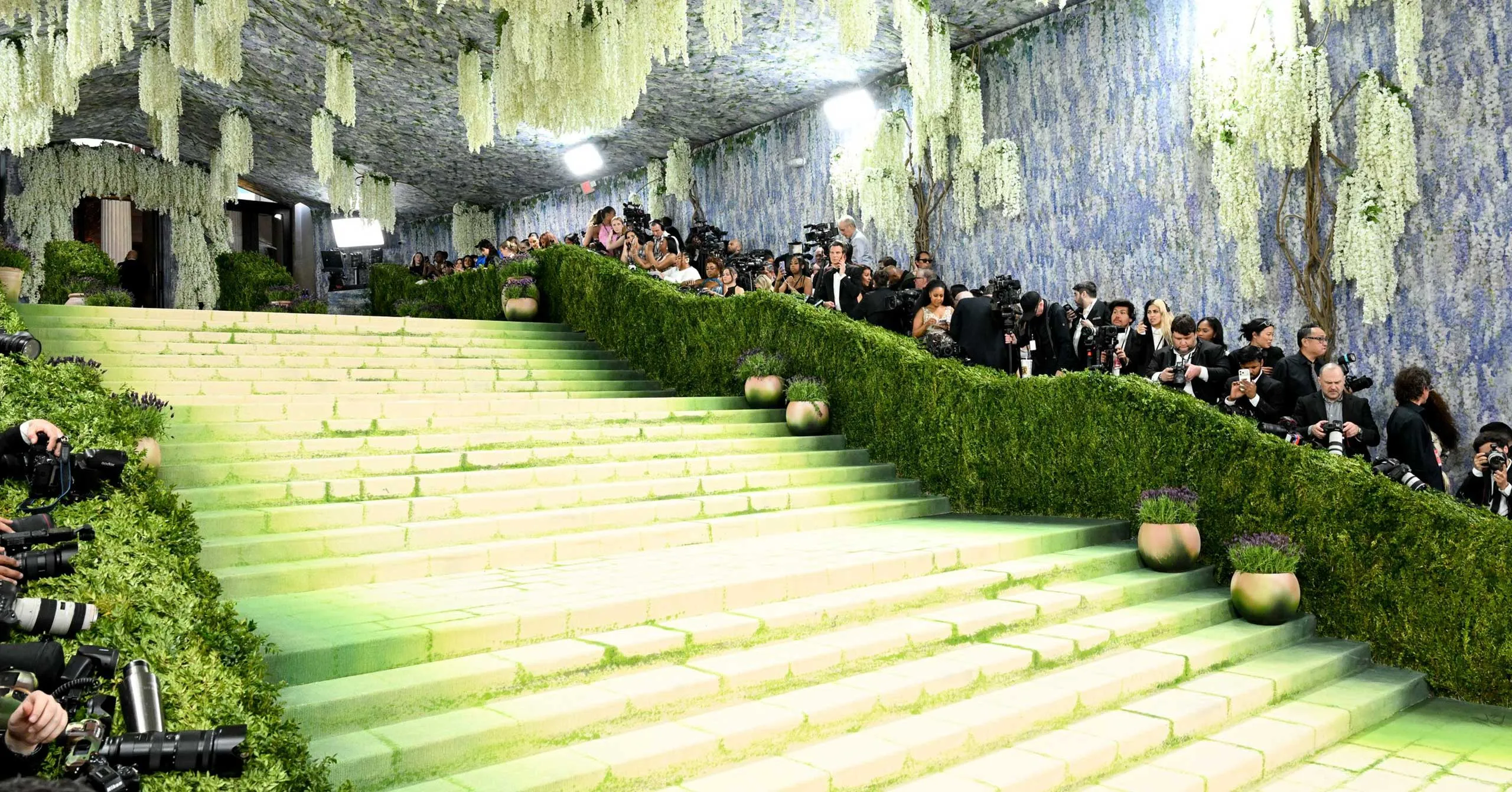 Green staircase at a fashion event with a line of photographers along the hedged walkway and hanging white flowers above.