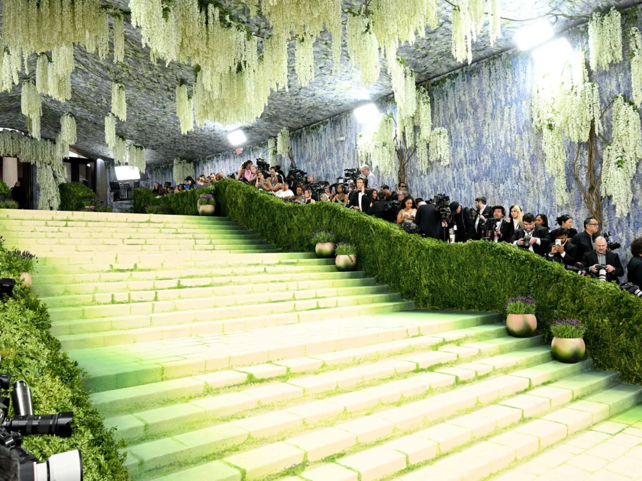 Green staircase at a fashion event with a line of photographers along the hedged walkway and hanging white flowers above.