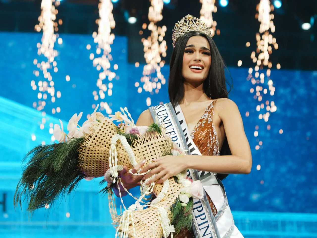 Beauty pageant winner wearing a crown and sash, holding a bouquet on stage with sparkling lights in the background.