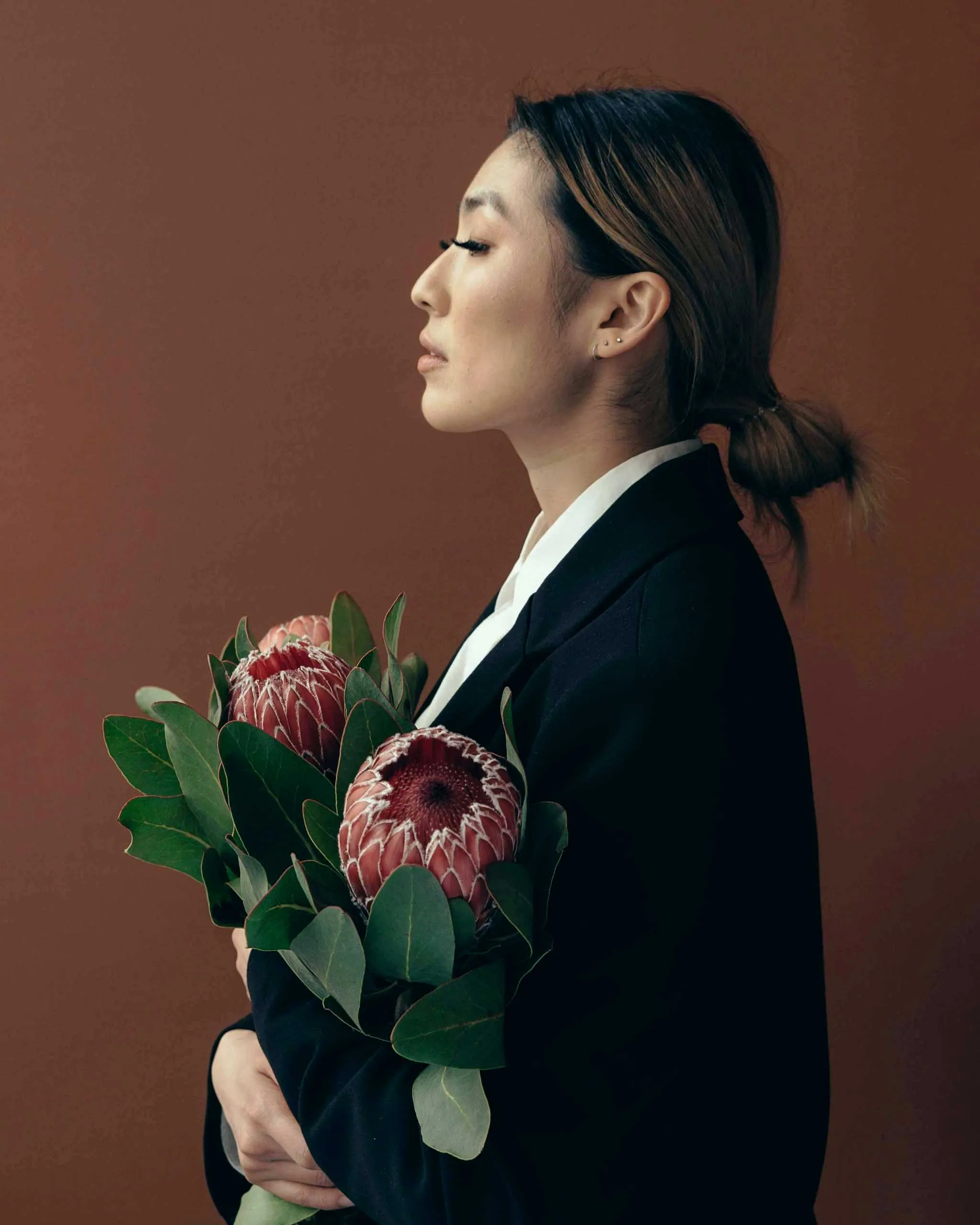 Profile view of a person in a dark blazer holding a bouquet of pink protea flowers against a brown background.