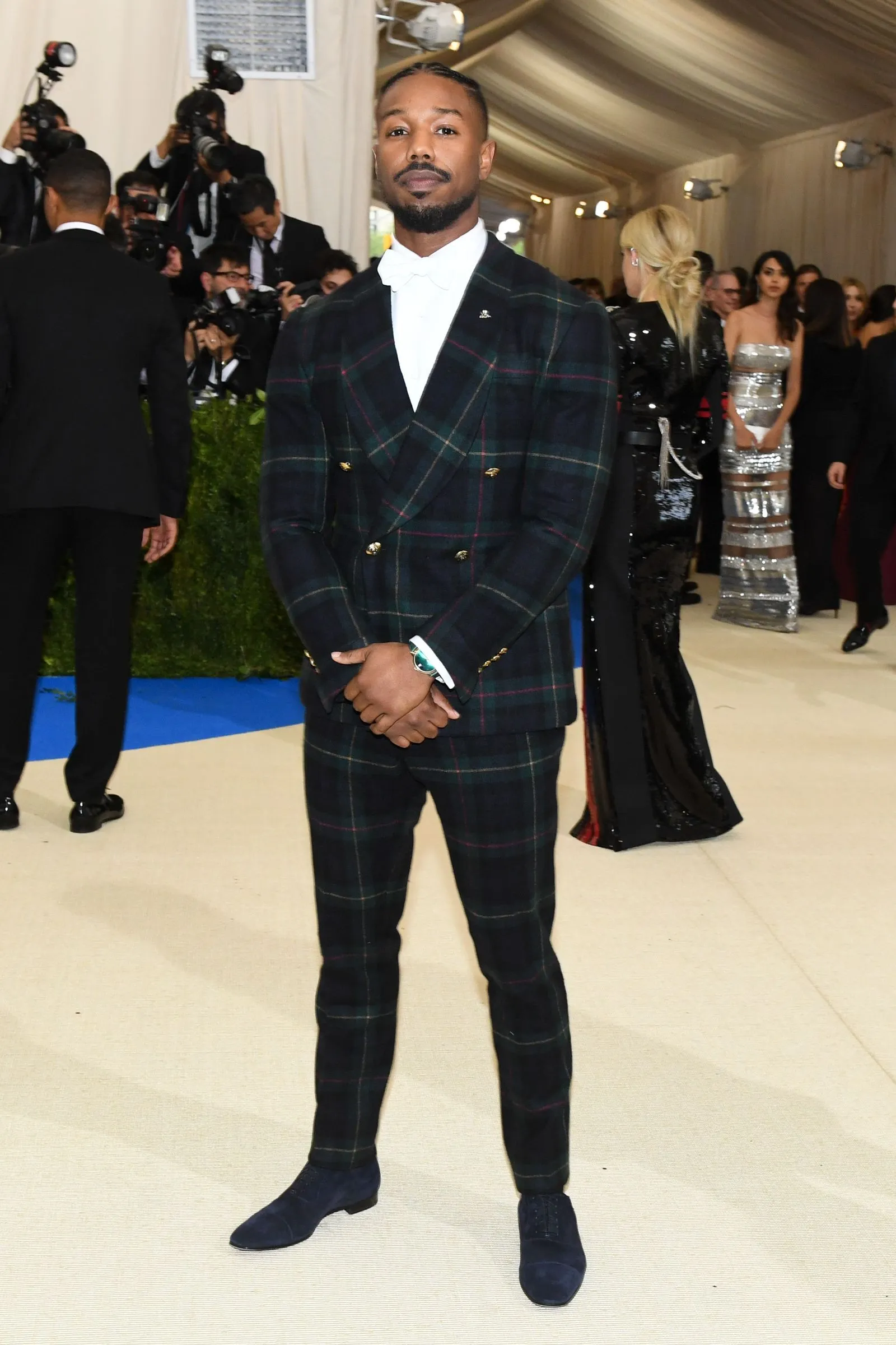 Man posing on a light-colored carpet in a dark green plaid double-breasted suit with a white shirt, hands clasped in front; photographers and guests in background.
