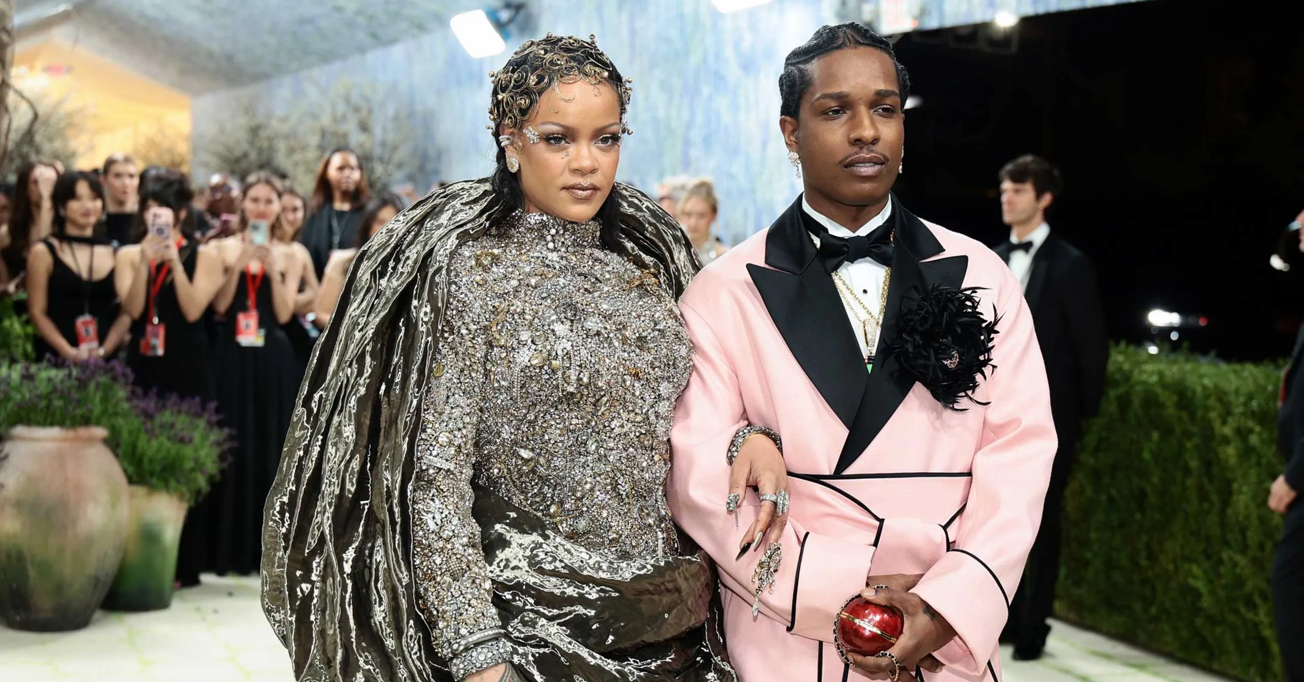 Two celebrities walk arm-in-arm in gala attire: a silver-embellished gown and a pink suit, as fans watch in the background.