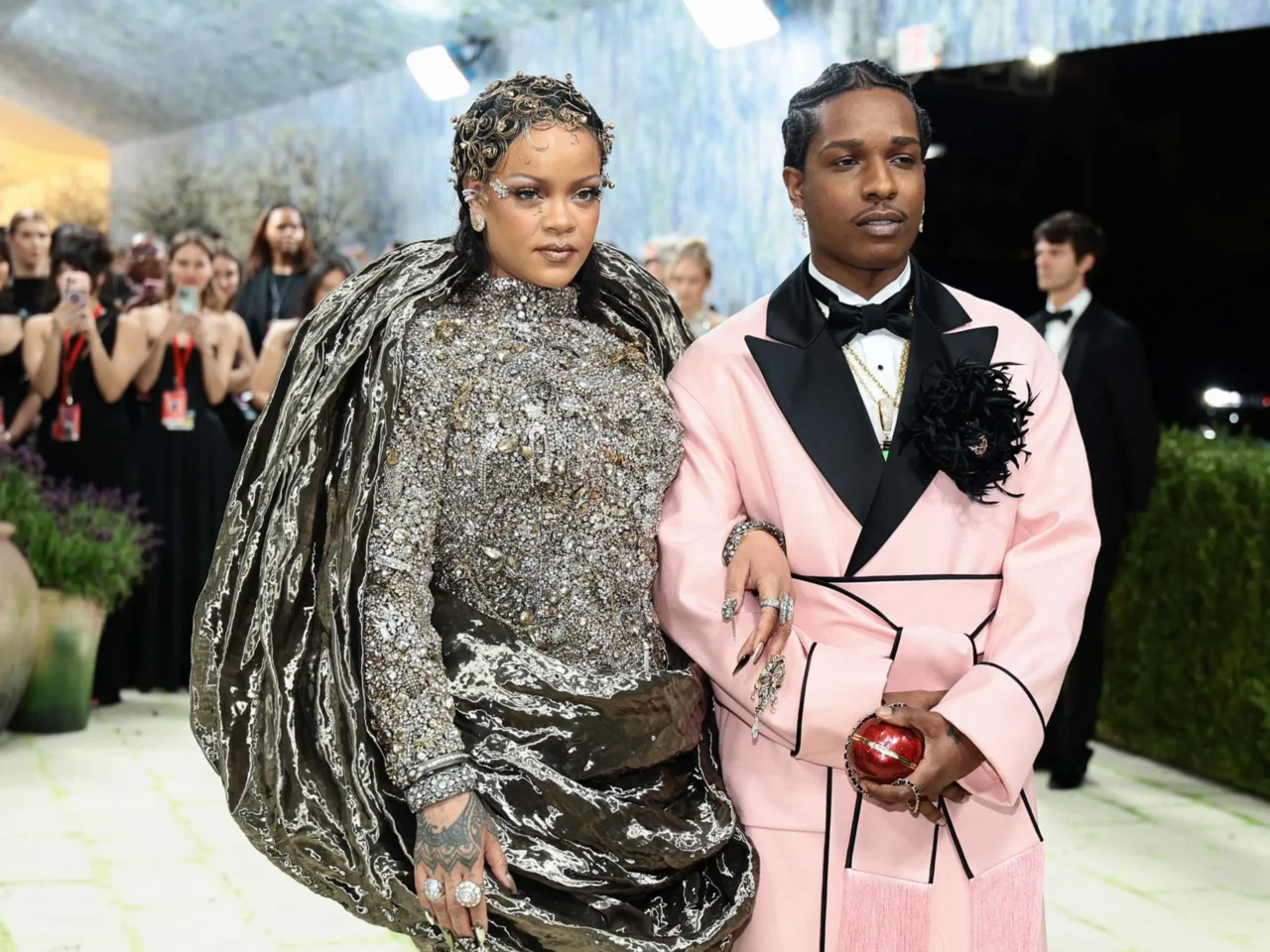 Two celebrities walk arm-in-arm in gala attire: a silver-embellished gown and a pink suit, as fans watch in the background.