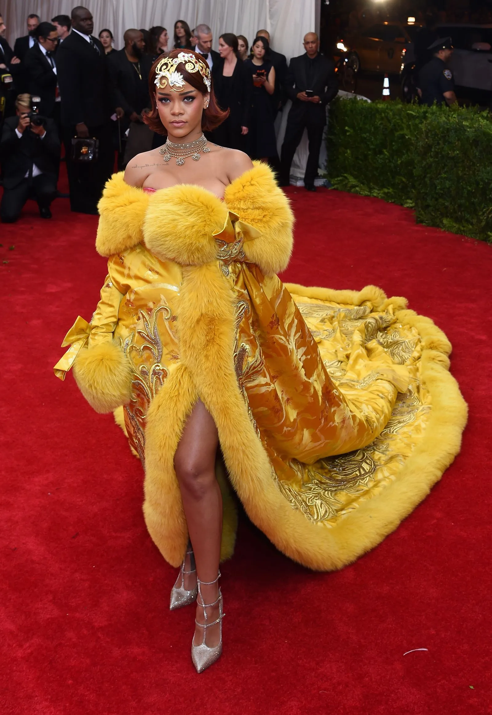 Celebrity on the red carpet wearing a gold-yellow, fur-trimmed gown with an ornate headpiece and diamond necklace, posing for photos.