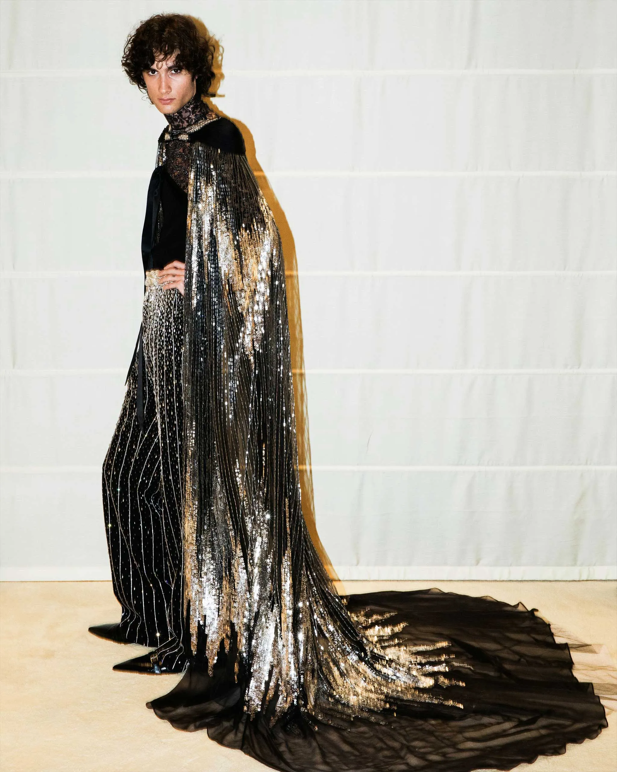 Person in a floor-length black and gold sequined gown with a dramatic cape, posed against a white backdrop.