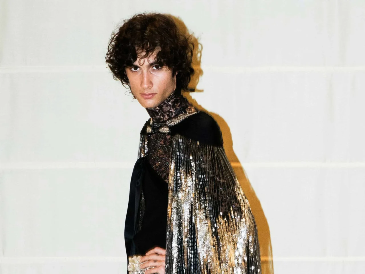 Portrait of a young man with dark curly hair wearing a black, fringed metallic top, looking at the camera against a pale backdrop.