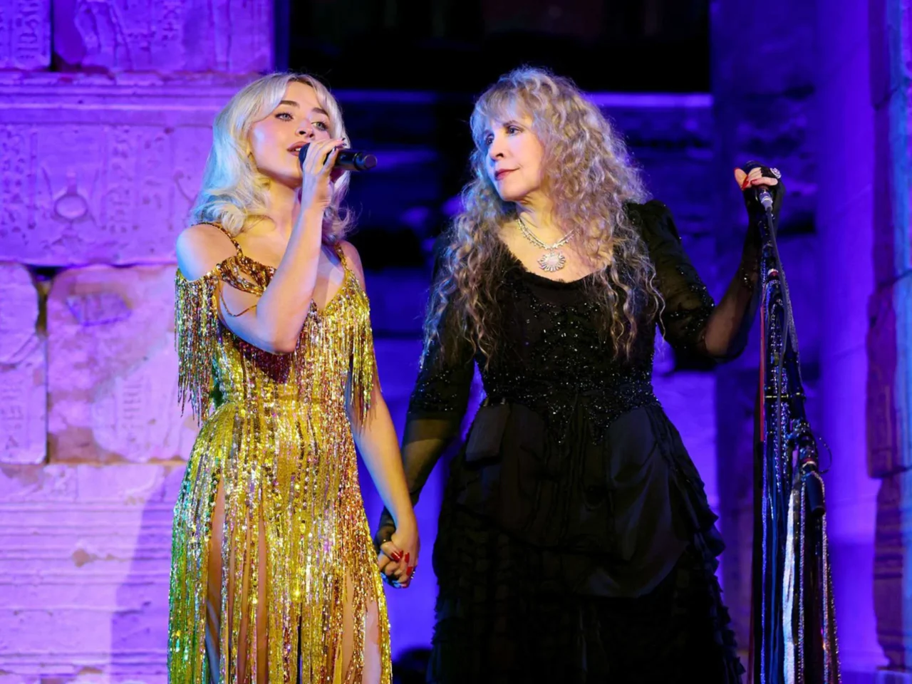 Two female singers perform on a stage with purple lighting; left singer wears a gold fringe dress and sings into a handheld microphone, right singer wears a black sequined outfit and sings into a microphone with a scarf.