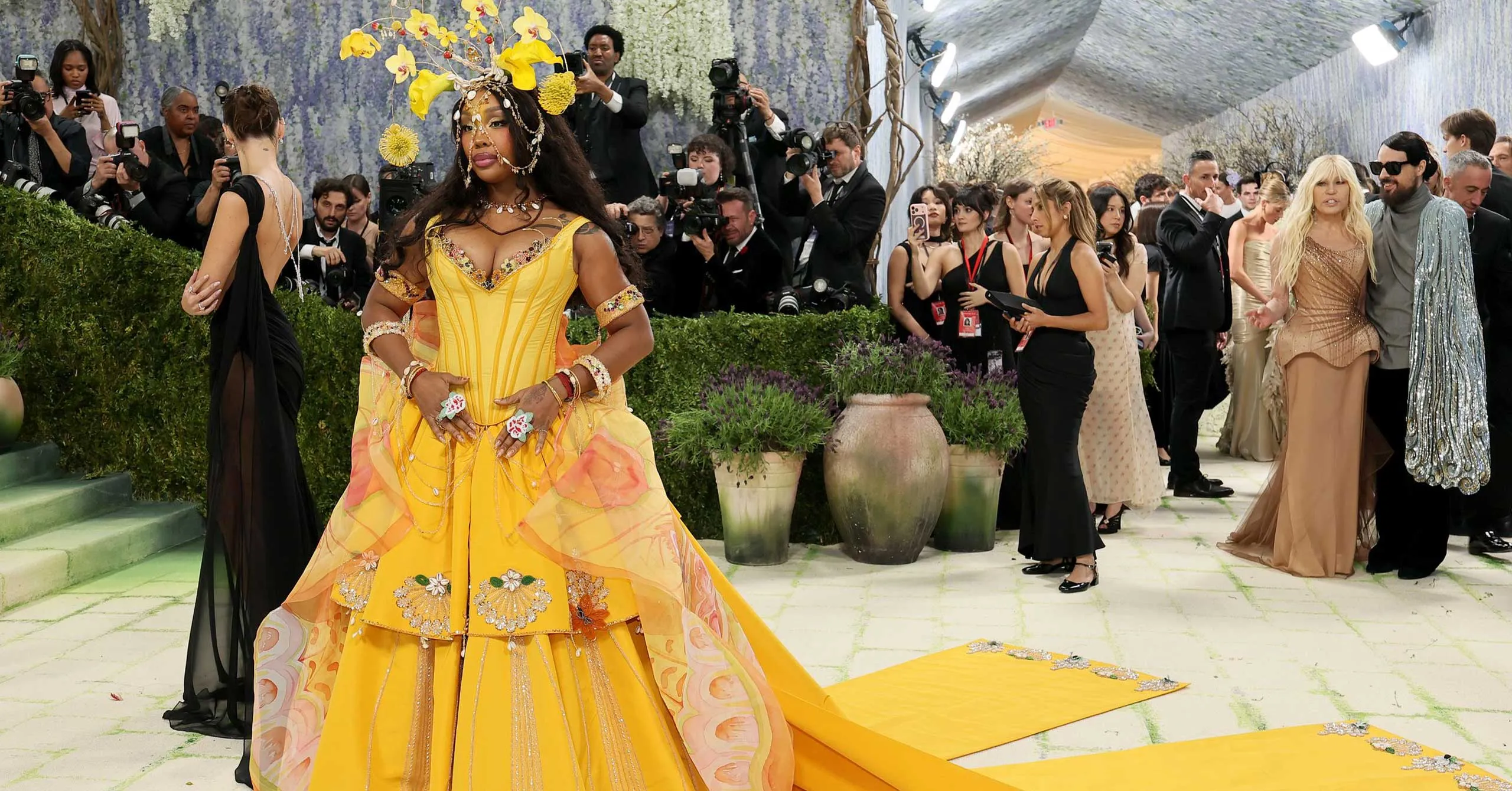 Woman in a bright yellow, ornate gown posing on a walkway at a red-carpet event, surrounded by photographers and onlookers.