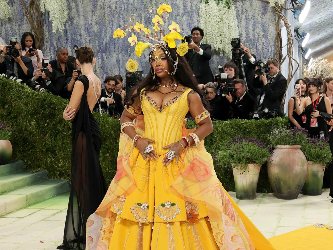 Woman in a bright yellow, ornate gown posing on a walkway at a red-carpet event, surrounded by photographers and onlookers.