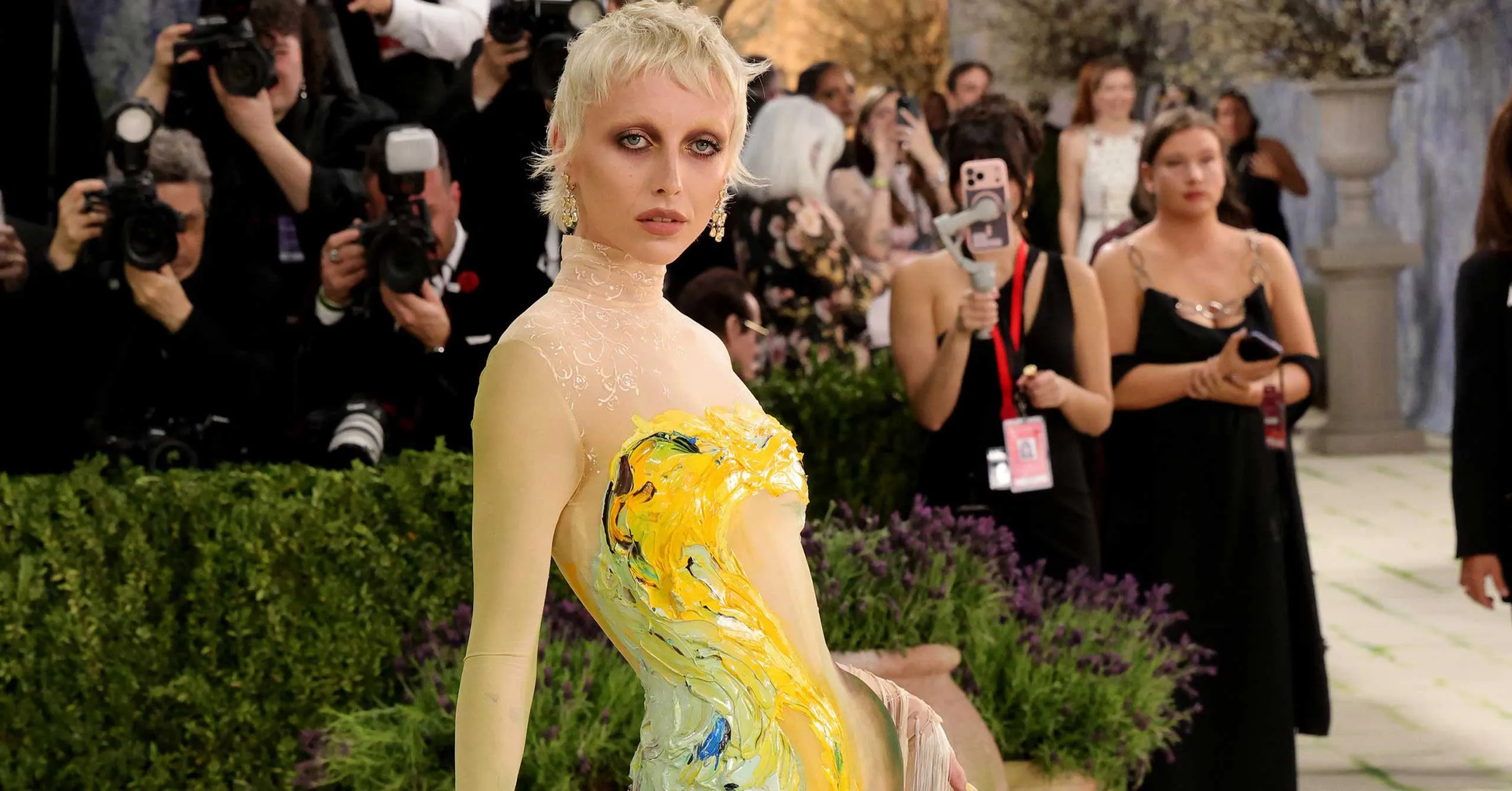 Model on a red carpet wearing a form-fitting gown with bold multicolor paint strokes; photographers crowd behind.