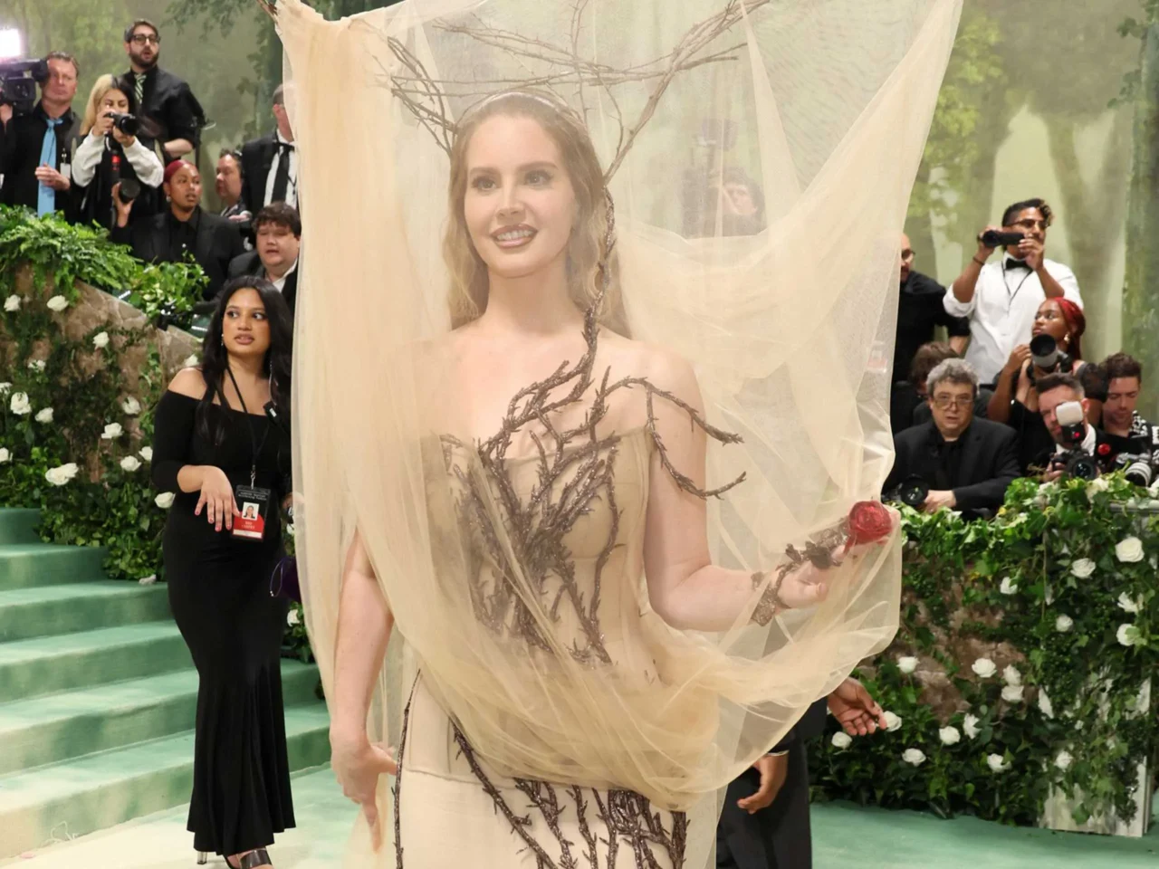 Model in a dramatic off-shoulder black gown with a sweeping train and a large pastel flower headpiece, posing on a green carpet as photographers line the backdrop.