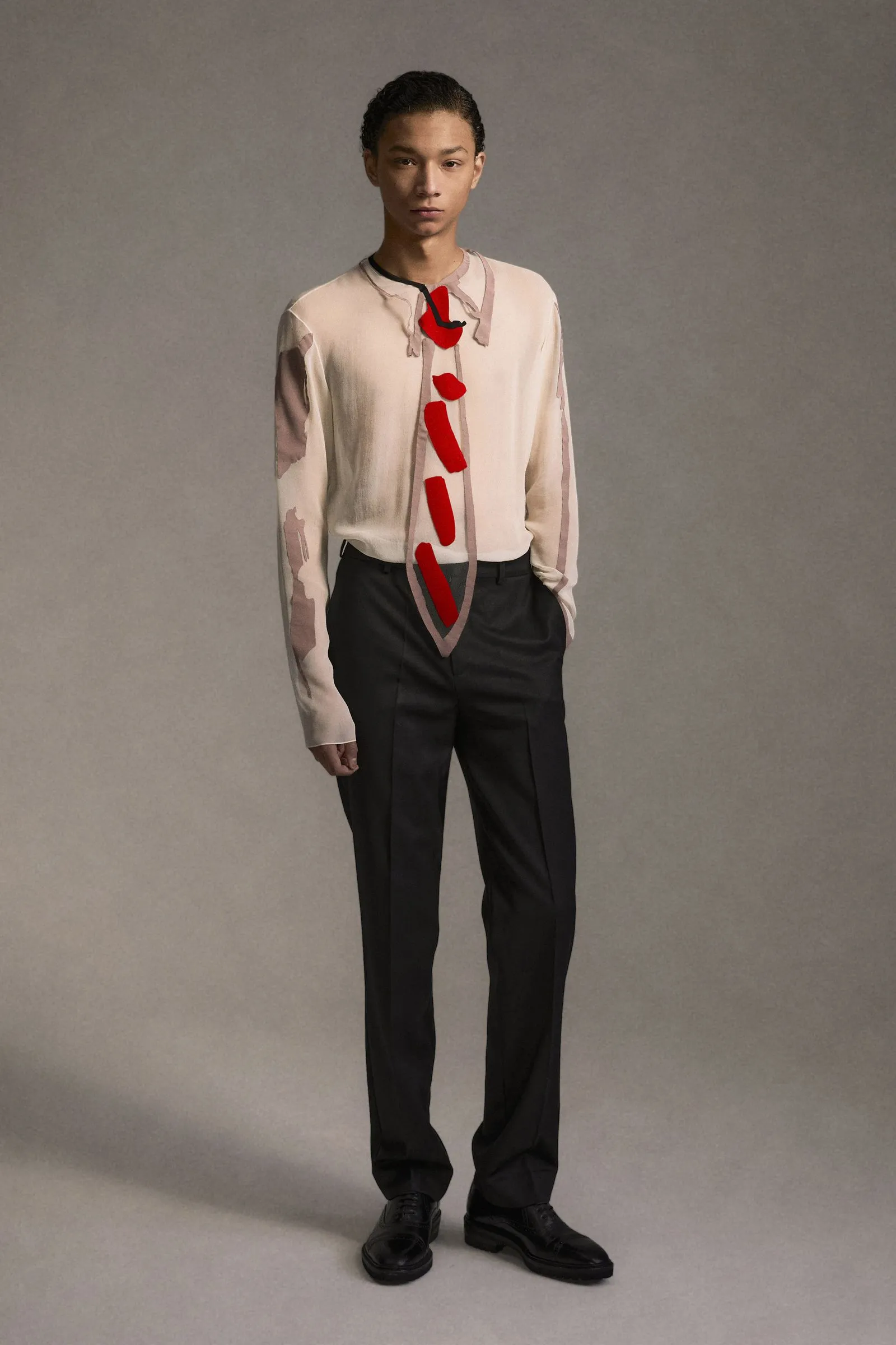Model standing in a neutral studio wearing a cream long-sleeve top with red abstract appliqué down the front, paired with black trousers and black shoes.