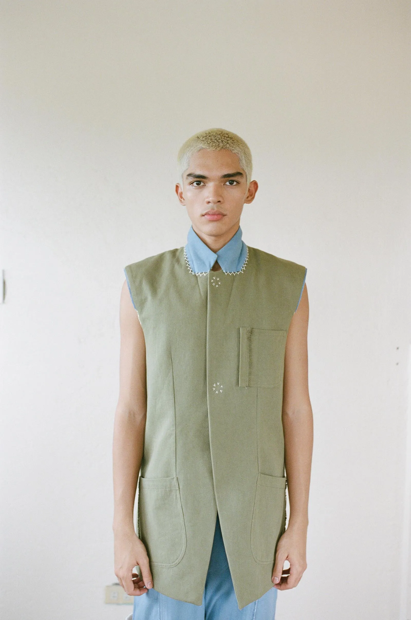 Person standing against a pale wall wearing an olive sleeveless vest over a blue collared shirt with a front pocket tag visible on the vest.