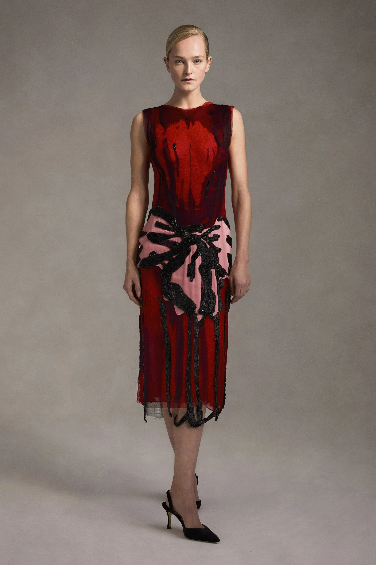Full-body fashion shot of a woman in a red sleeveless dress with black fringe and a pink panel, standing against a neutral backdrop in high heels.