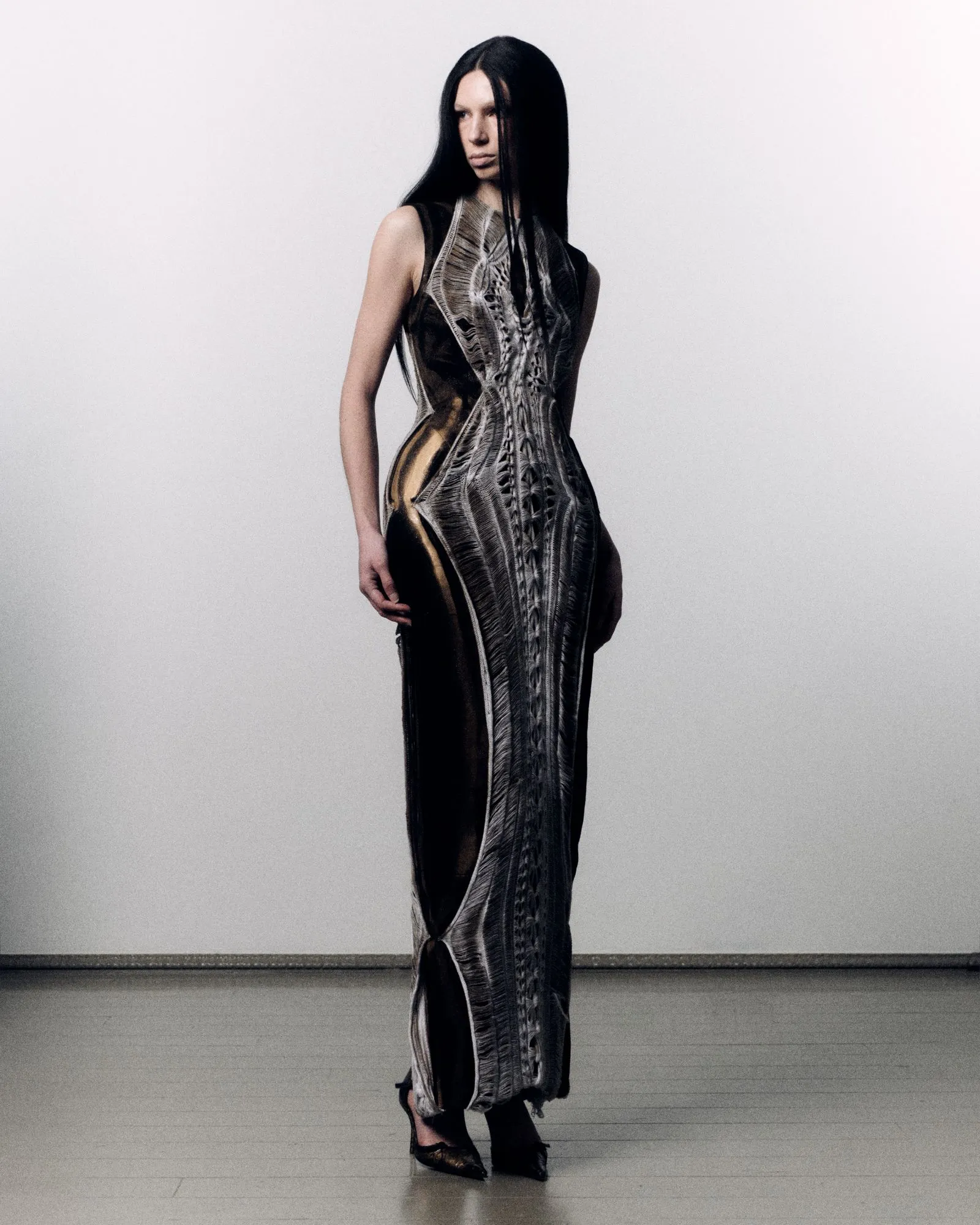 Model wearing a long metallic-patterned evening gown with black hair, standing against a pale studio backdrop.