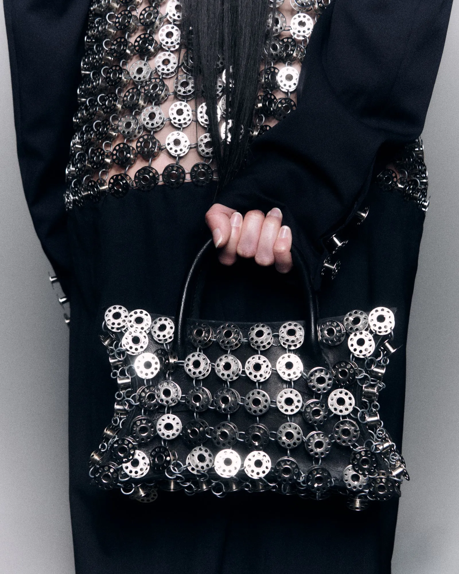 Close-up of a black garment densely covered in silver circular metal discs and chains, with a hand resting near the waist area.