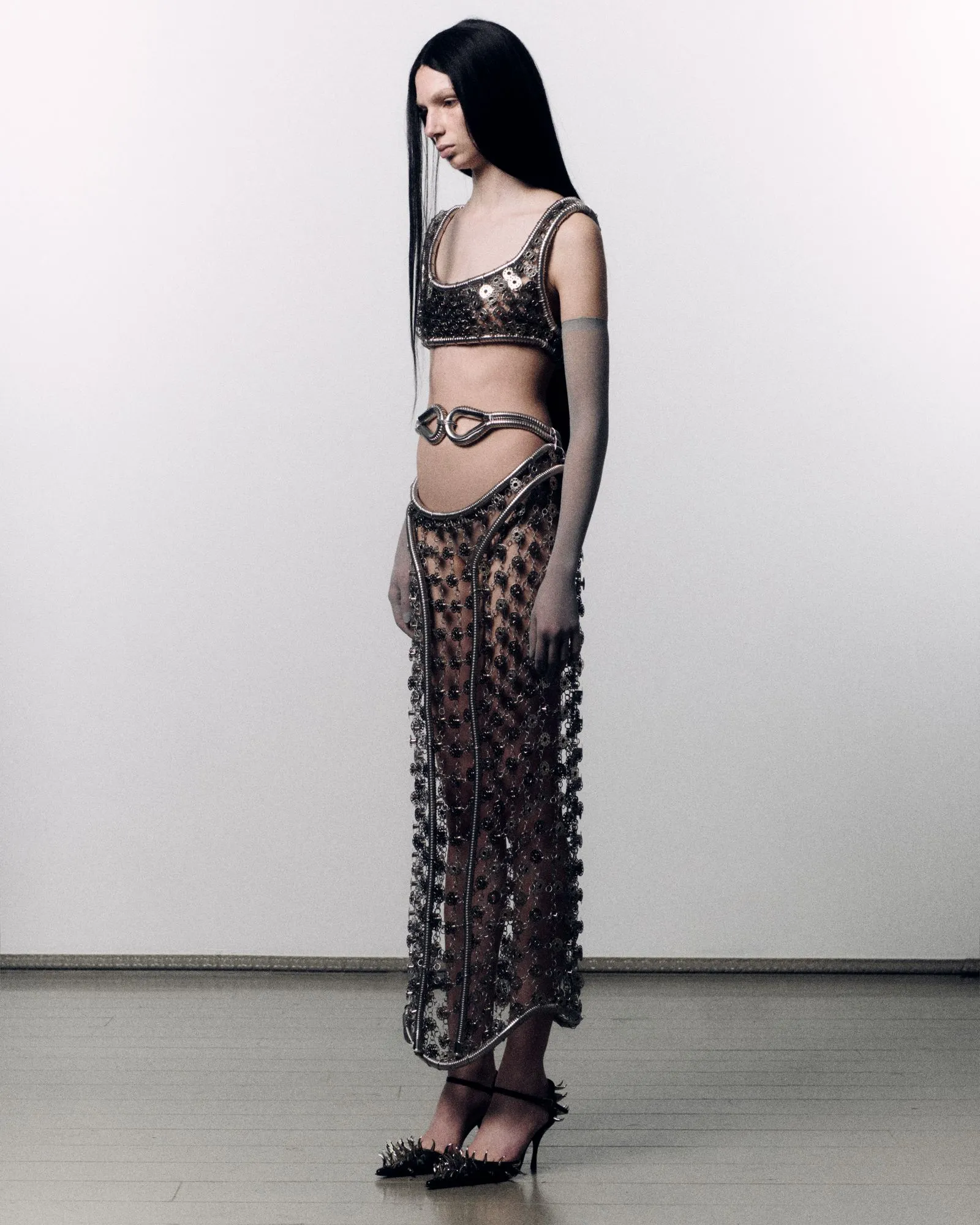 Profile view of a model wearing a metallic beaded crop top and sheer, chain-draped skirt with spiked high heels.