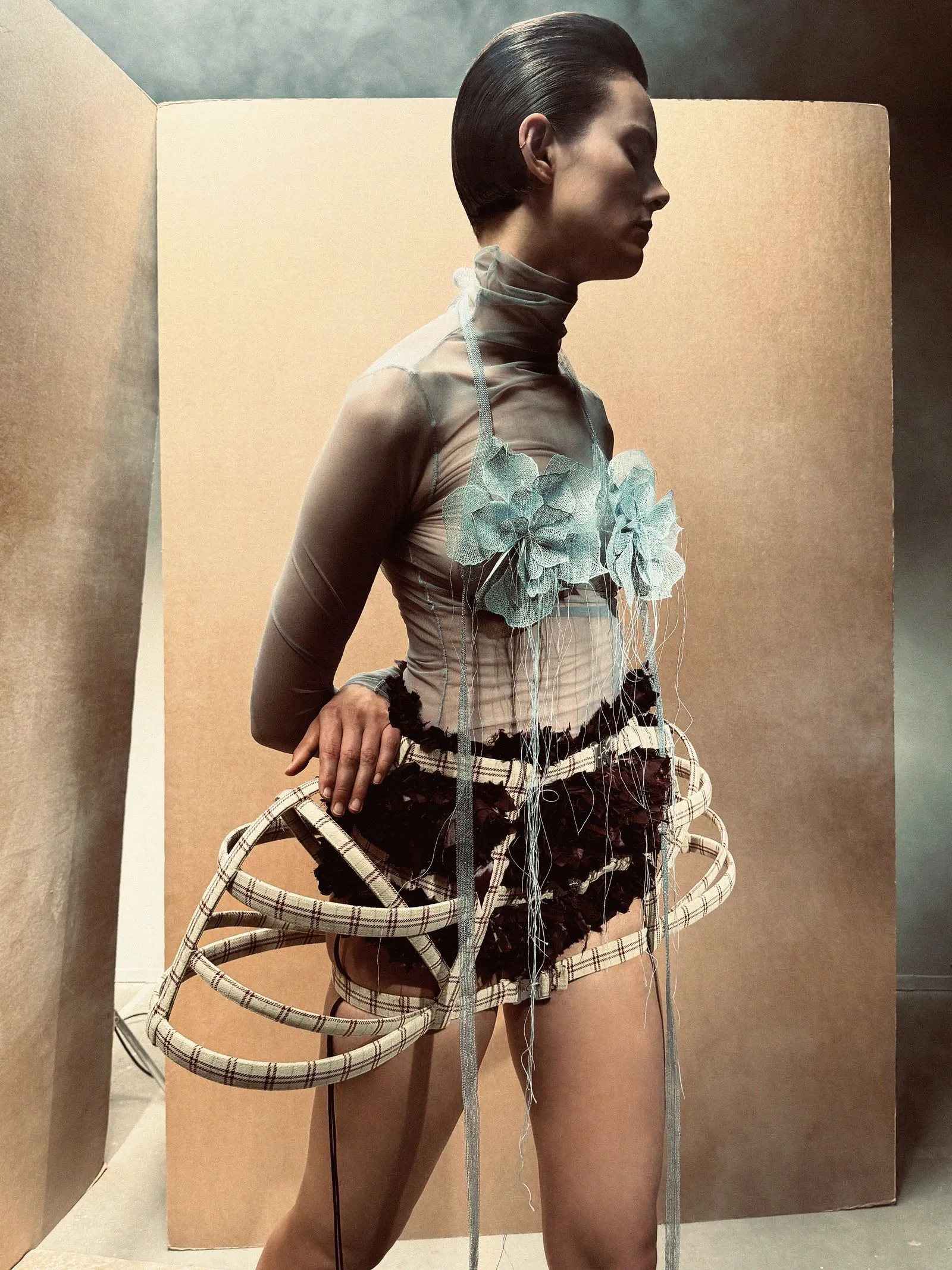 Avant-garde fashion portrait: model in a sheer top with blue fabric flowers and long ribbons, wearing a plaid skirt, posed beside cardboard panels.