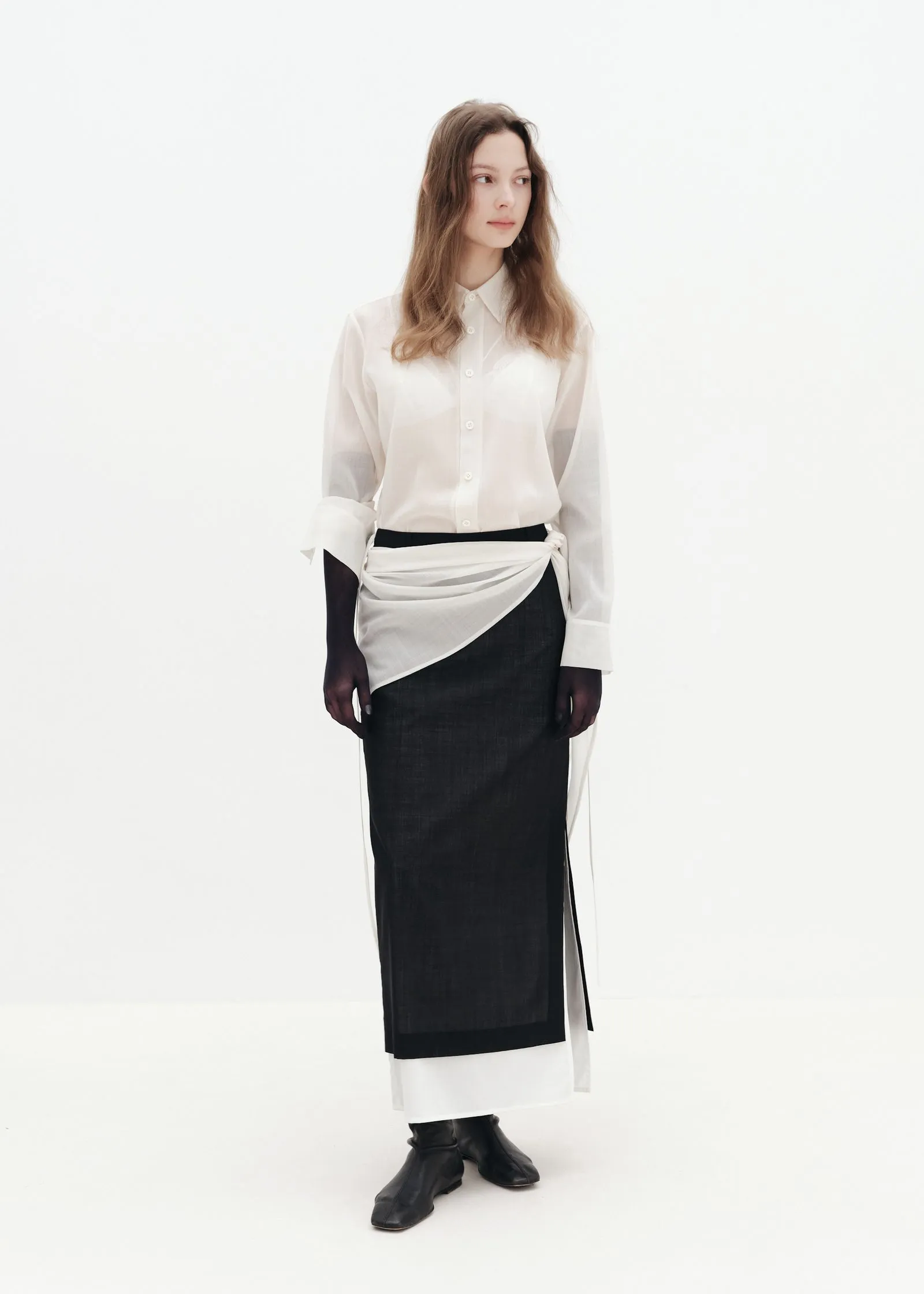 Model standing in a white studio background, wearing a white button-down shirt with a black long skirt and a white wrap sash at the waist, looking slightly to the right.