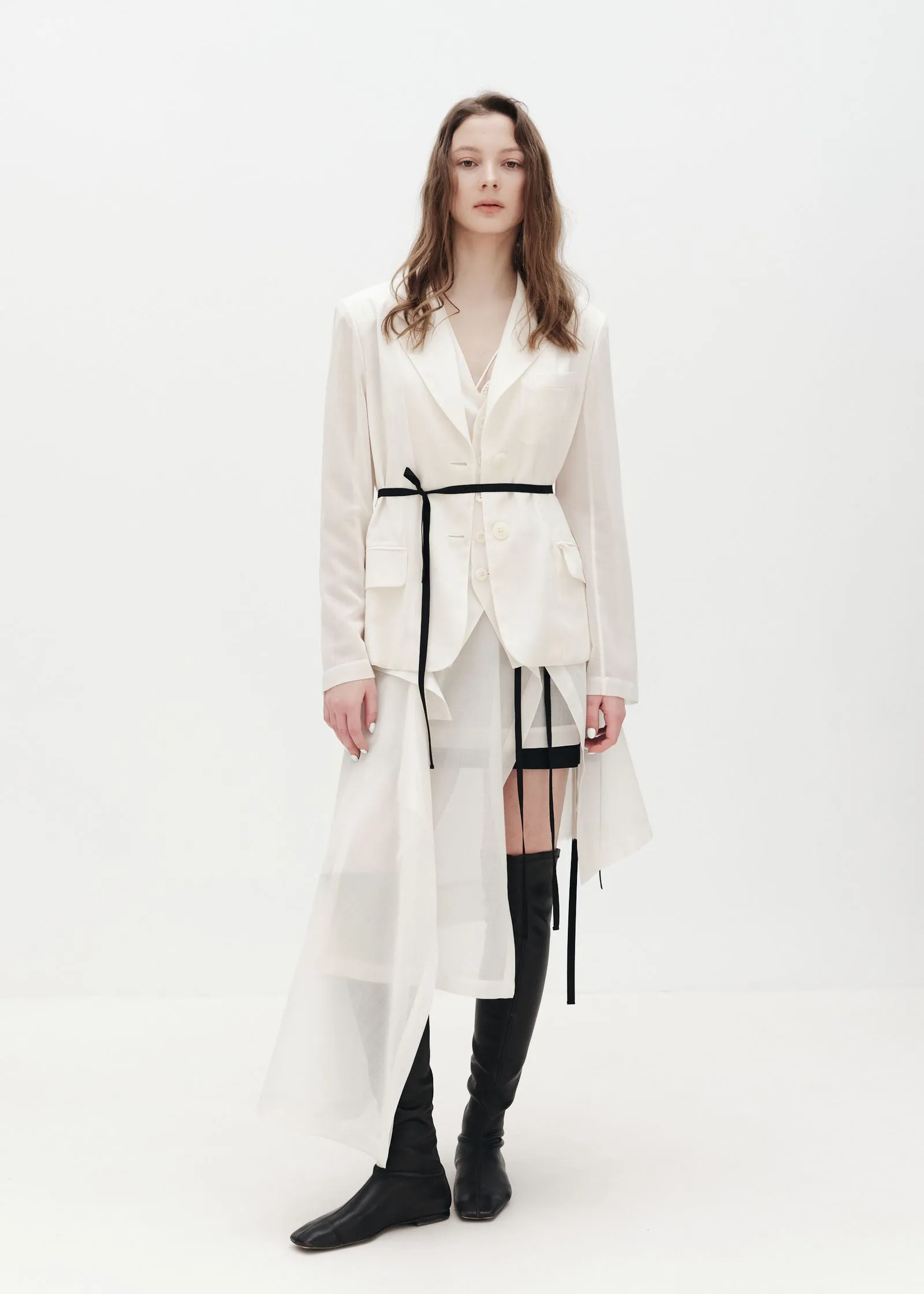 Model wearing a white tailored blazer and matching skirt with a black belt, standing against a white backdrop in a minimalist fashion pose.