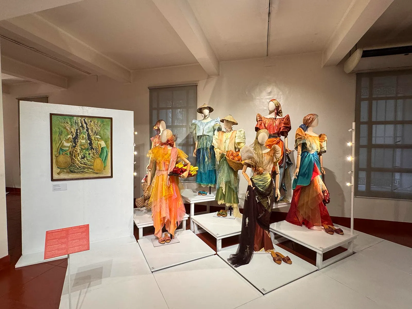 Museum display of colorful vintage dresses on mannequins arranged on white platforms, with a framed painting on the left panel.