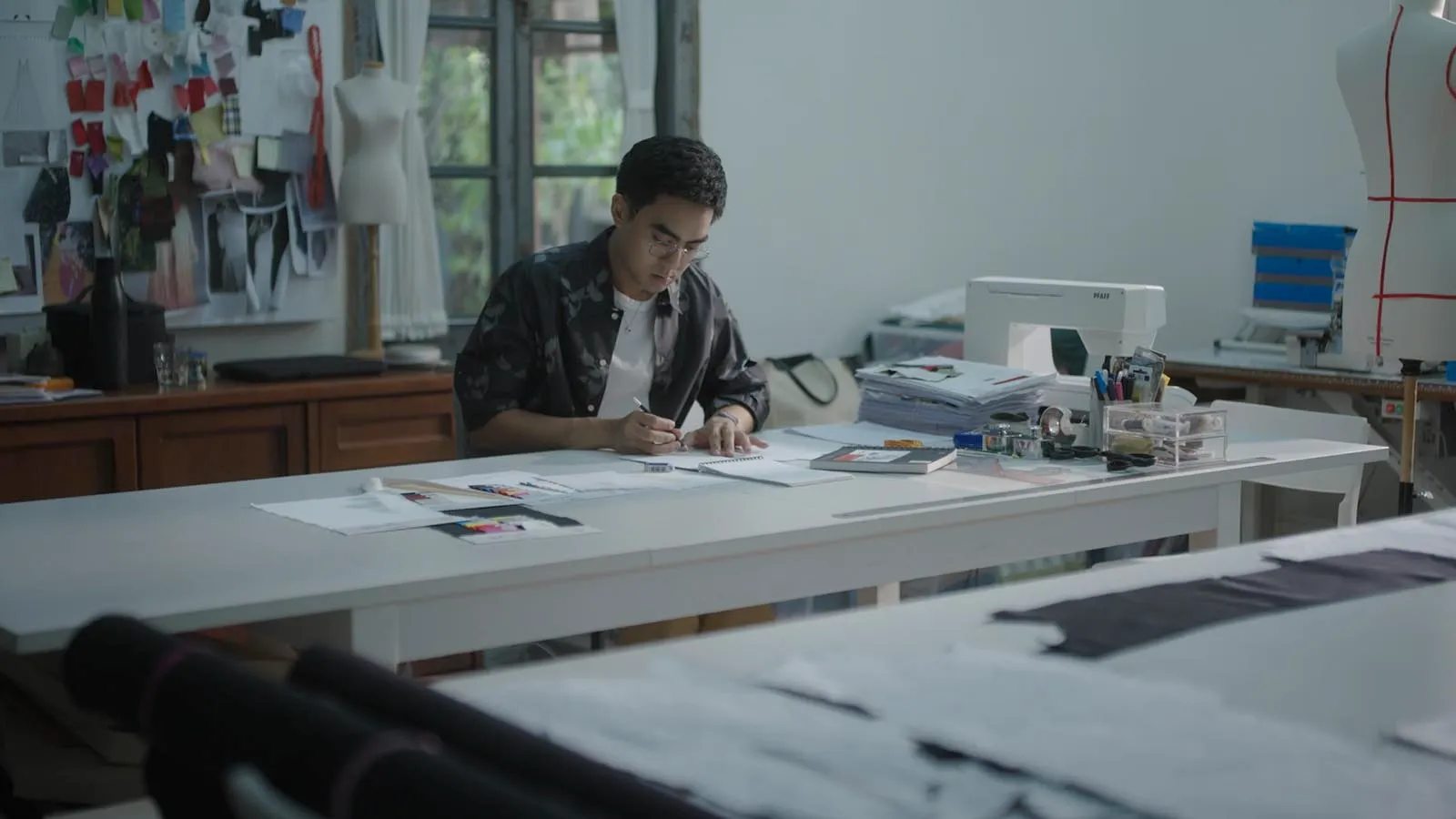Young fashion designer at a white workspace, drafting patterns with swatches and sewing tools nearby.