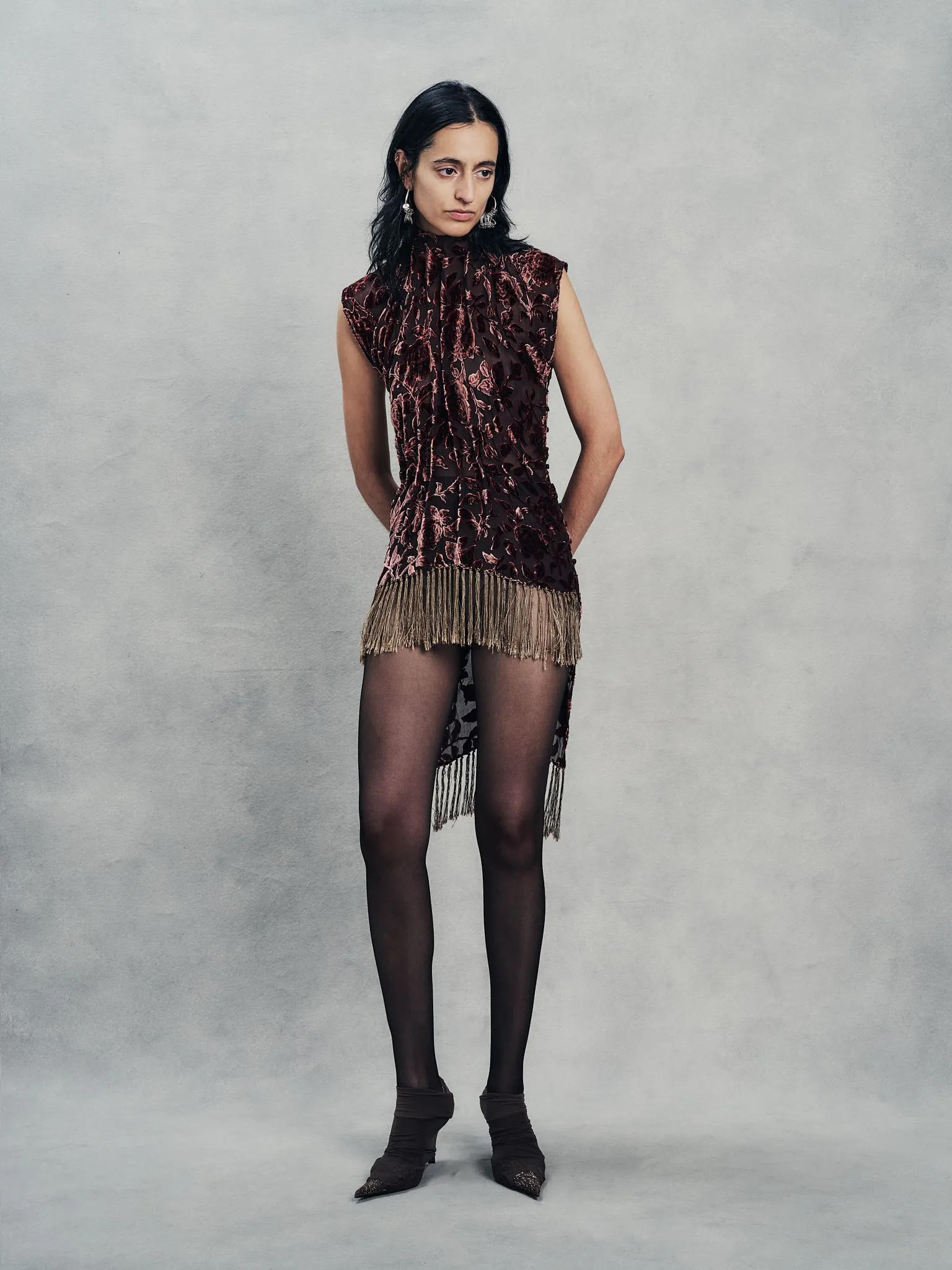 Model posing in a burgundy velvet dress with floral pattern and long fringe hemlines, standing with hands behind her, against a gray textured backdrop.
