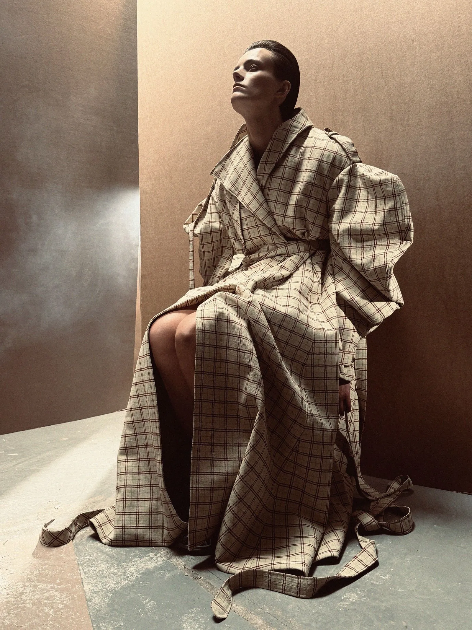 Person seated in a large beige plaid coat, looking upward against a textured brown wall.
