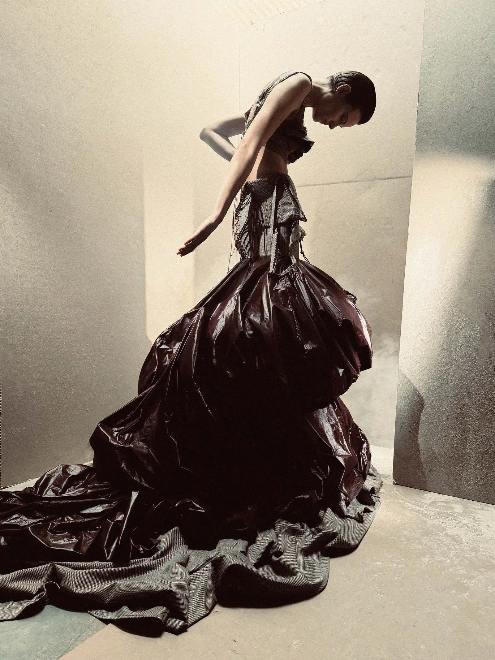 Person wearing an oversized black plastic bag dress, bending forward in a light-toned studio.